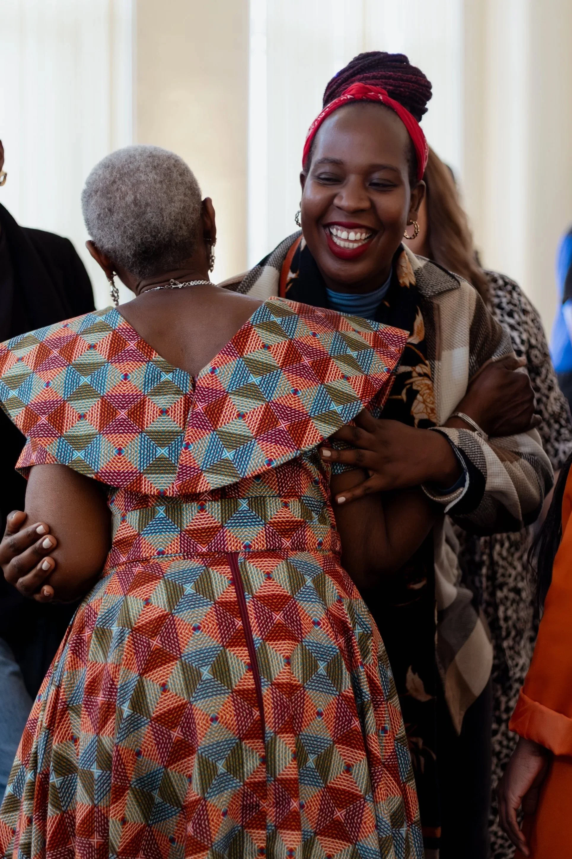 Two women smiling, embracing, and engaging in a friendly hug at an indoor social gathering. The woman facing away has short gray hair and wears a colorful, patterned dress, while the woman facing the camera has braided hair styled in a bun with a red headband, and wears a jacket with a floral shirt underneath.