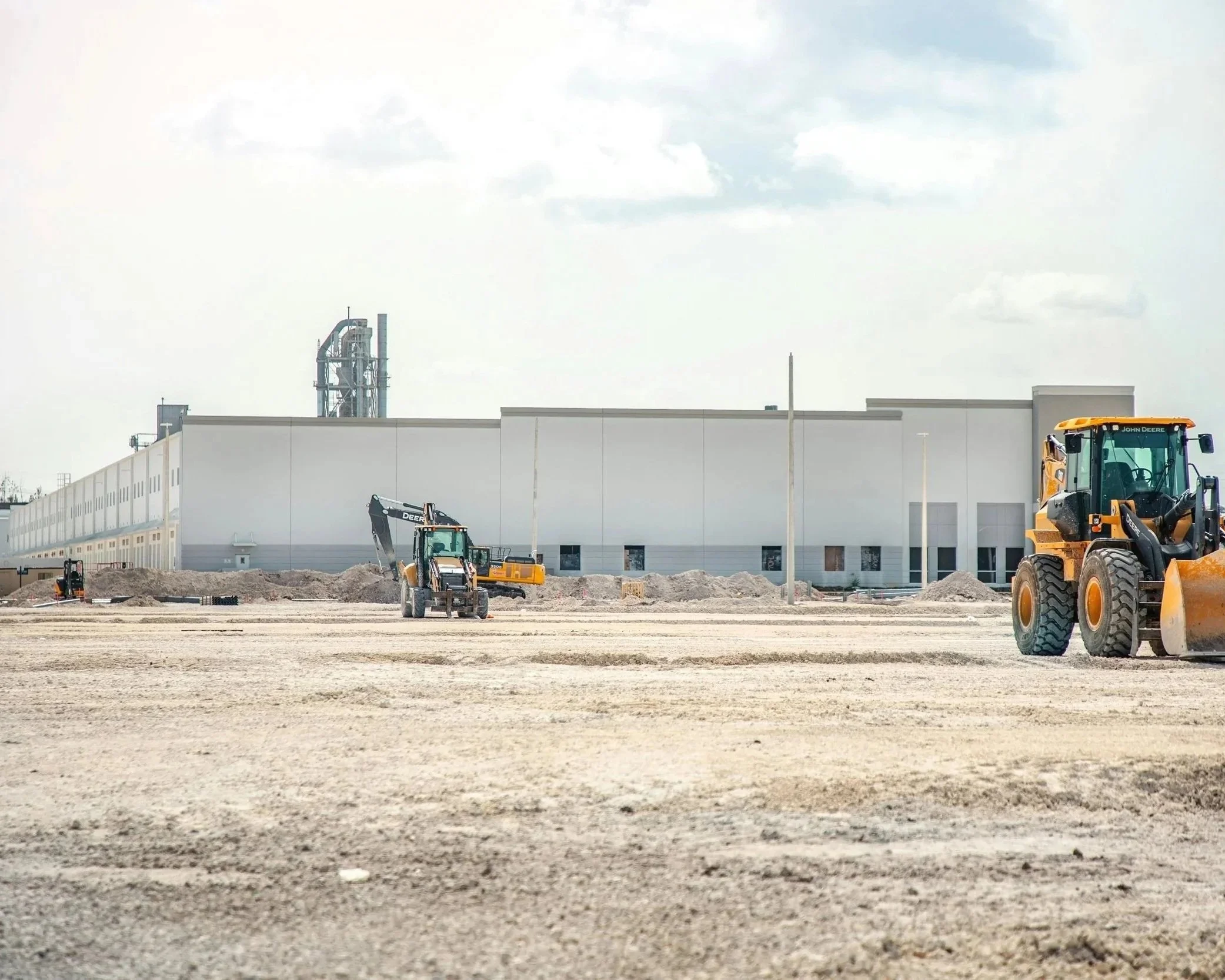 Chantier de construction avec des engins de terrassement, bâtiment blanc en arrière-plan, ciel nuageux.
