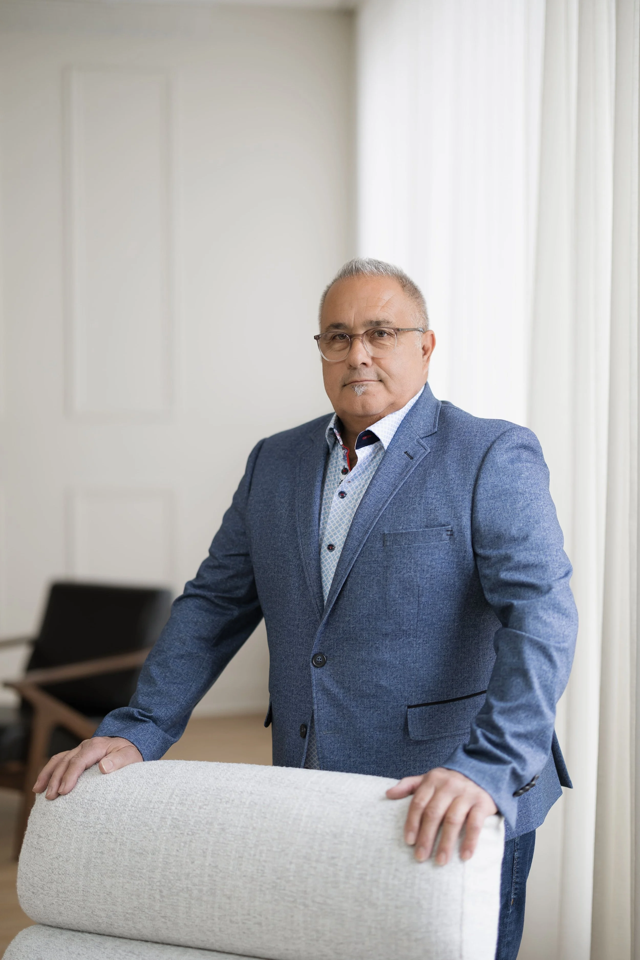 Homme d'affaires portant un costume bleu, cheveux gris, lunettes, dans une salle avec des rideaux blancs.