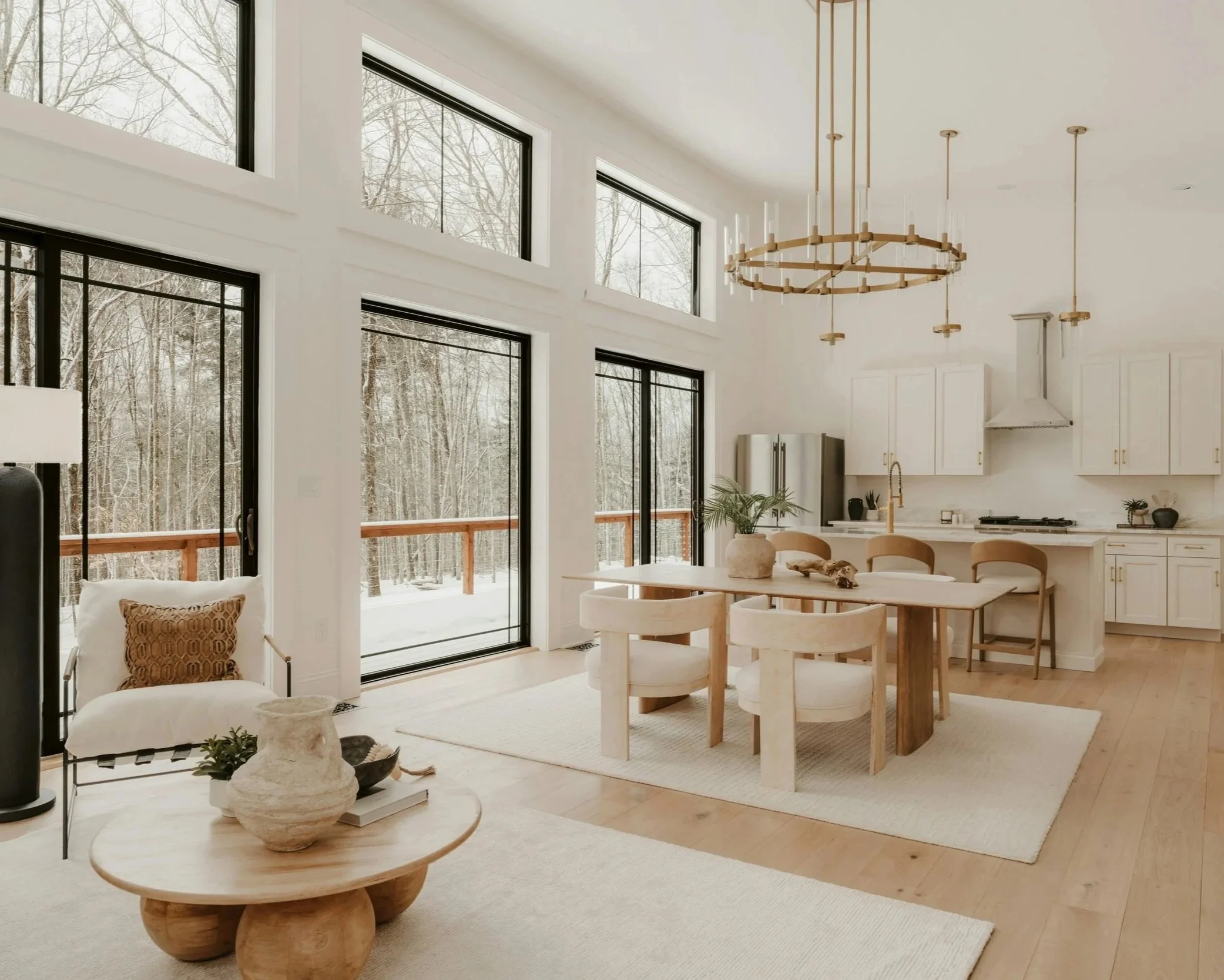 Intérieur d'une cuisine moderne avec de grandes fenêtres donnant sur un paysage enneigé, mobilier blanc, table en bois clair avec sept chaises, éclairage suspendu doré, coin détente avec chaise et table basse en bois, décor minimaliste et lumineux.