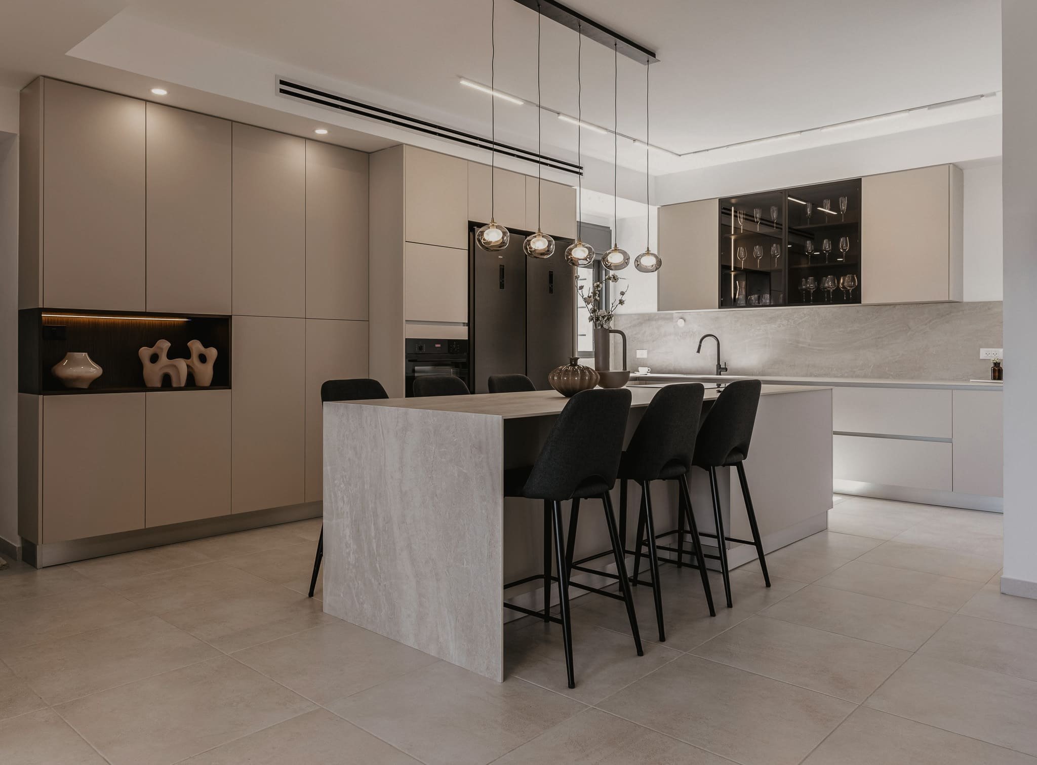 Cuisine moderne avec îlot central, meubles beige, étagères en verre avec vaisselle, éclairage suspendu, décor minimaliste avec vases et fleurs.