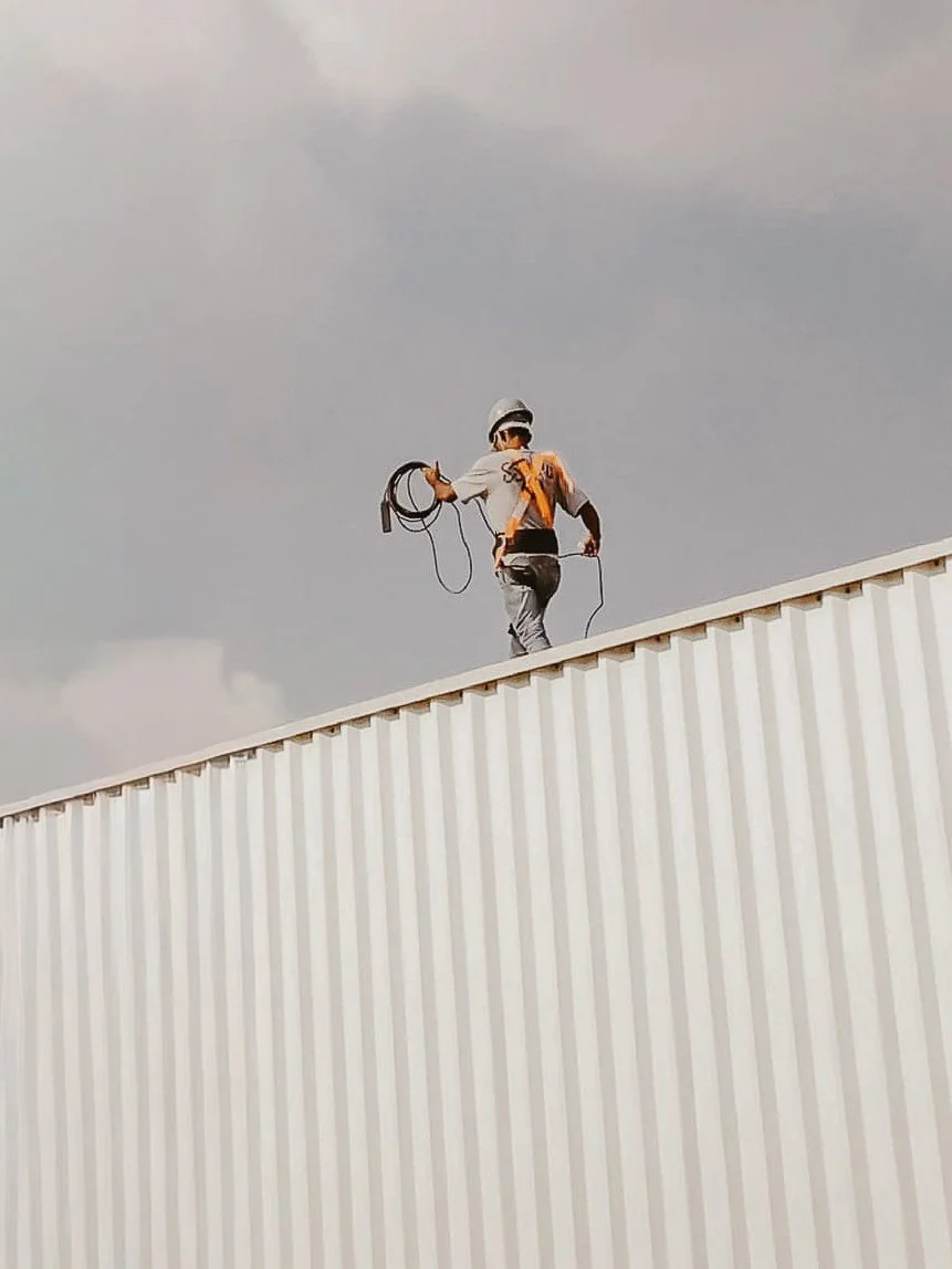 Un ouvrier en casque de sécurité et équipement de sécurité marche sur le toit d'un bâtiment industriel, tenant un câble dans les mains, sous un ciel nuageux.
