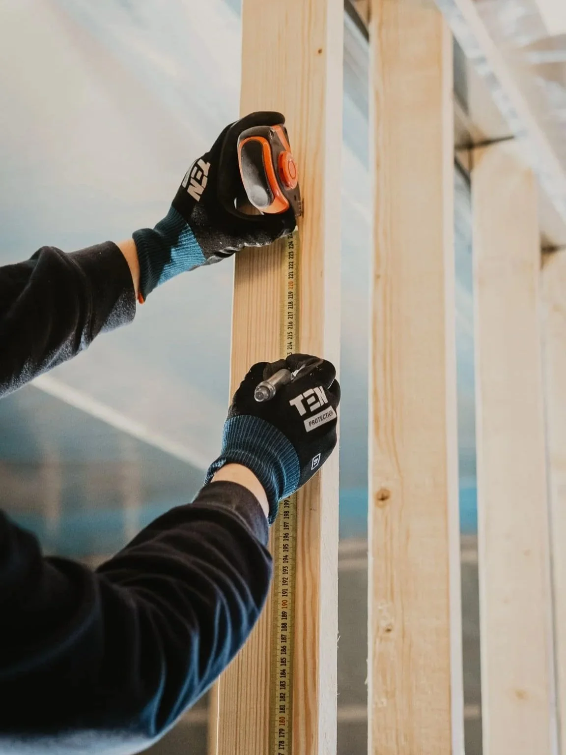 Une personne portant des gants noirs et bleus mesure une pièce en bois à l'aide d'une règle métallique lors de la construction d'une structure en bois.