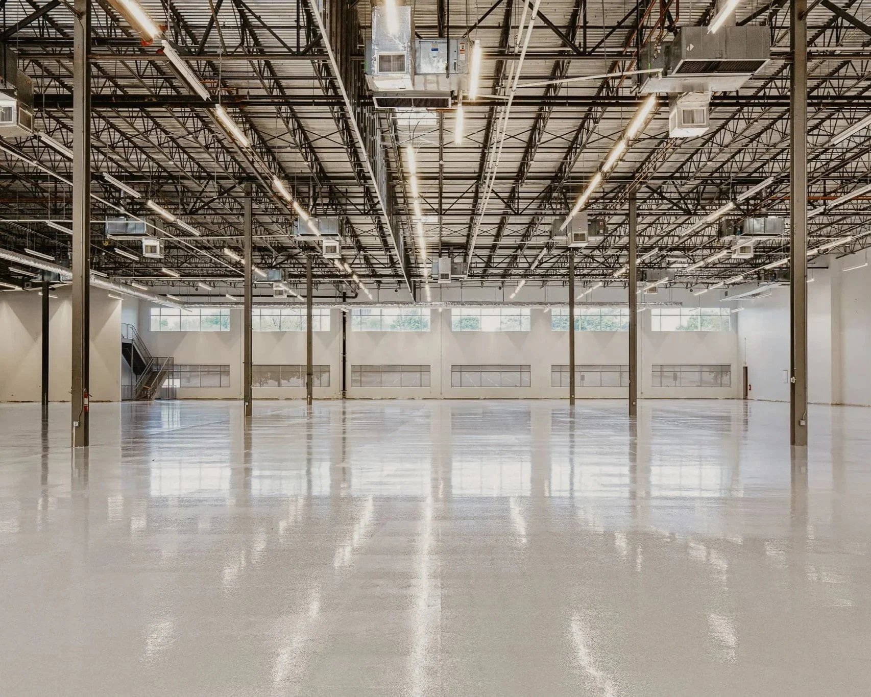 Intérieur d'un grand espace industriel ou une salle vide avec un sol brillant, des murs blancs, de grandes fenêtres et un plafond avec des luminaires suspendus et des conduits.