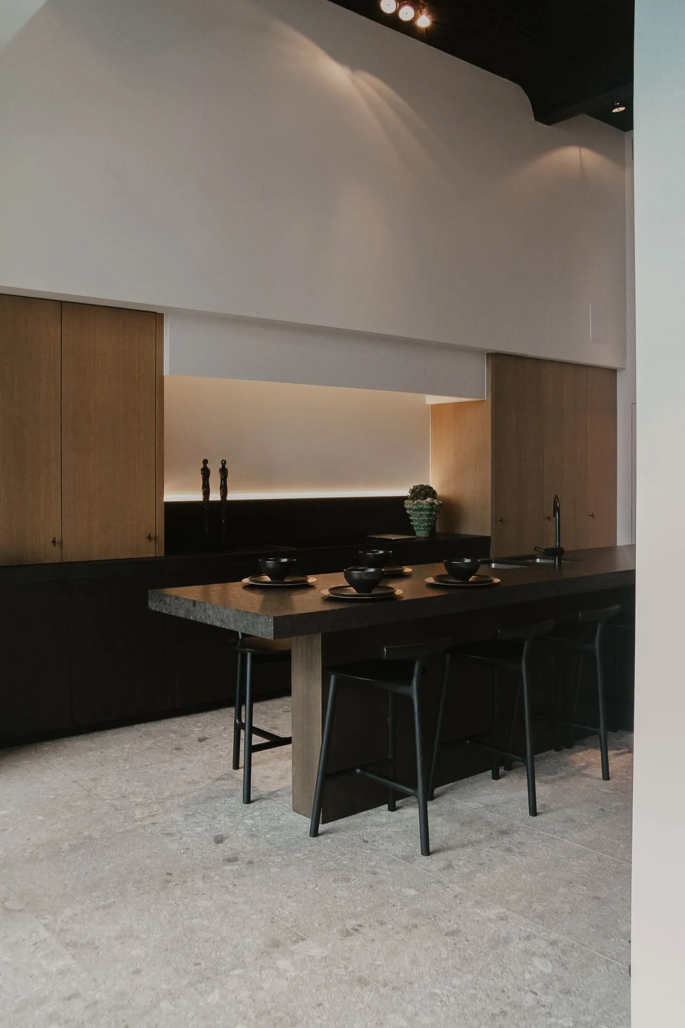 Cuisine moderne avec îlot noir, meubles en bois clair, assiettes et bols noirs sur l'îlot, décoration minimaliste avec deux sculptures et une plante verte.