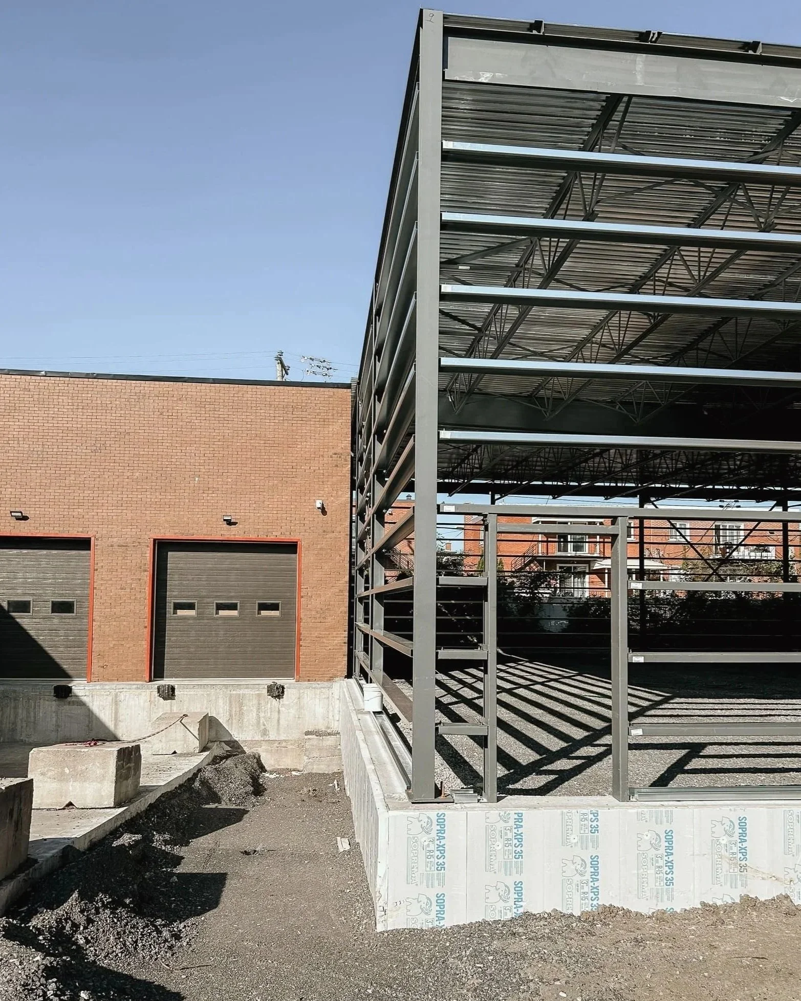 Vue d'une structure métallique en construction à côté d'un bâtiment en brique, avec deux portes de garage, sous un ciel bleu.