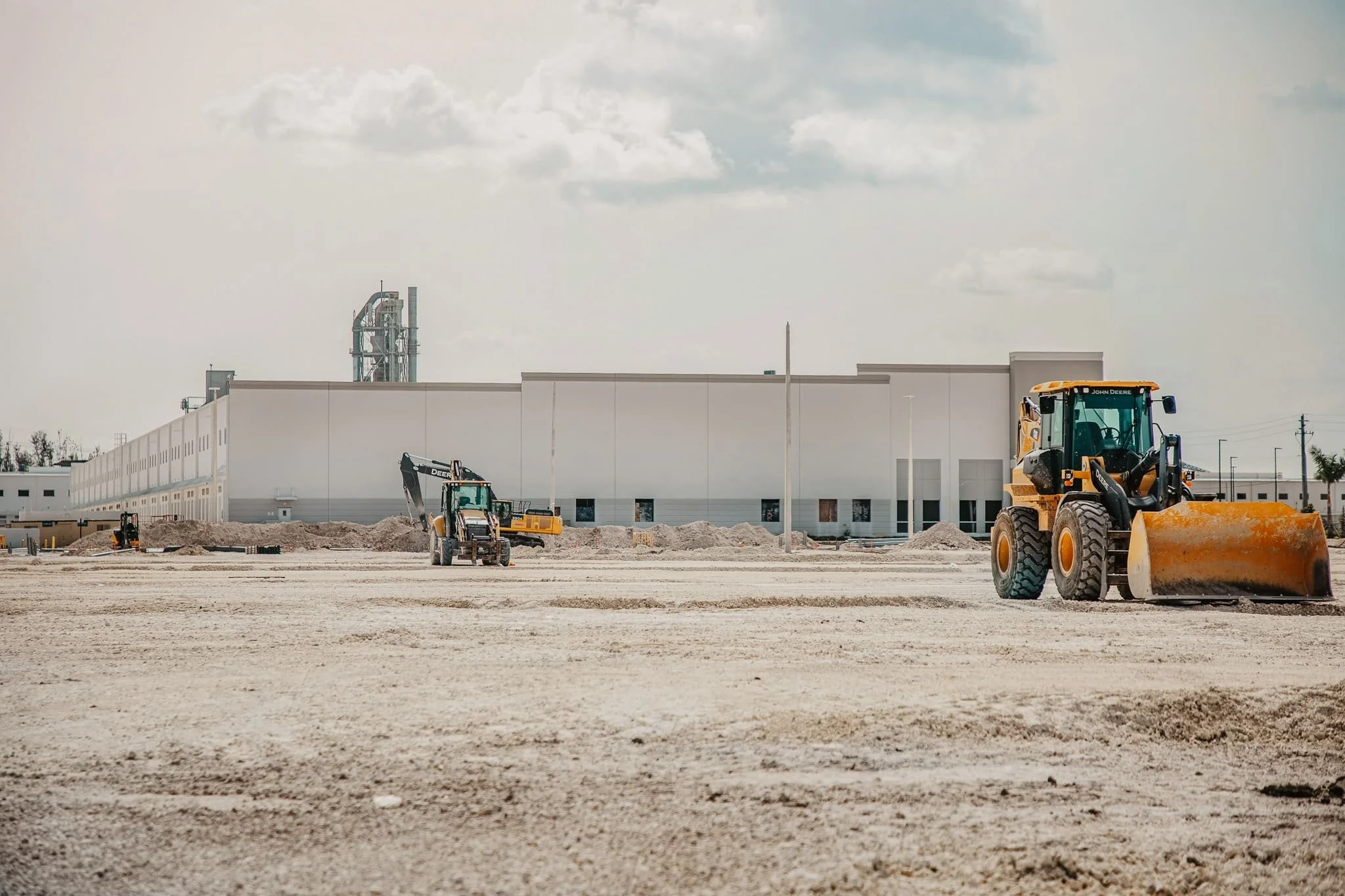 Chantier de construction avec des engins de terrassement en action devant un bâtiment blanc, sous un ciel nuageux.
