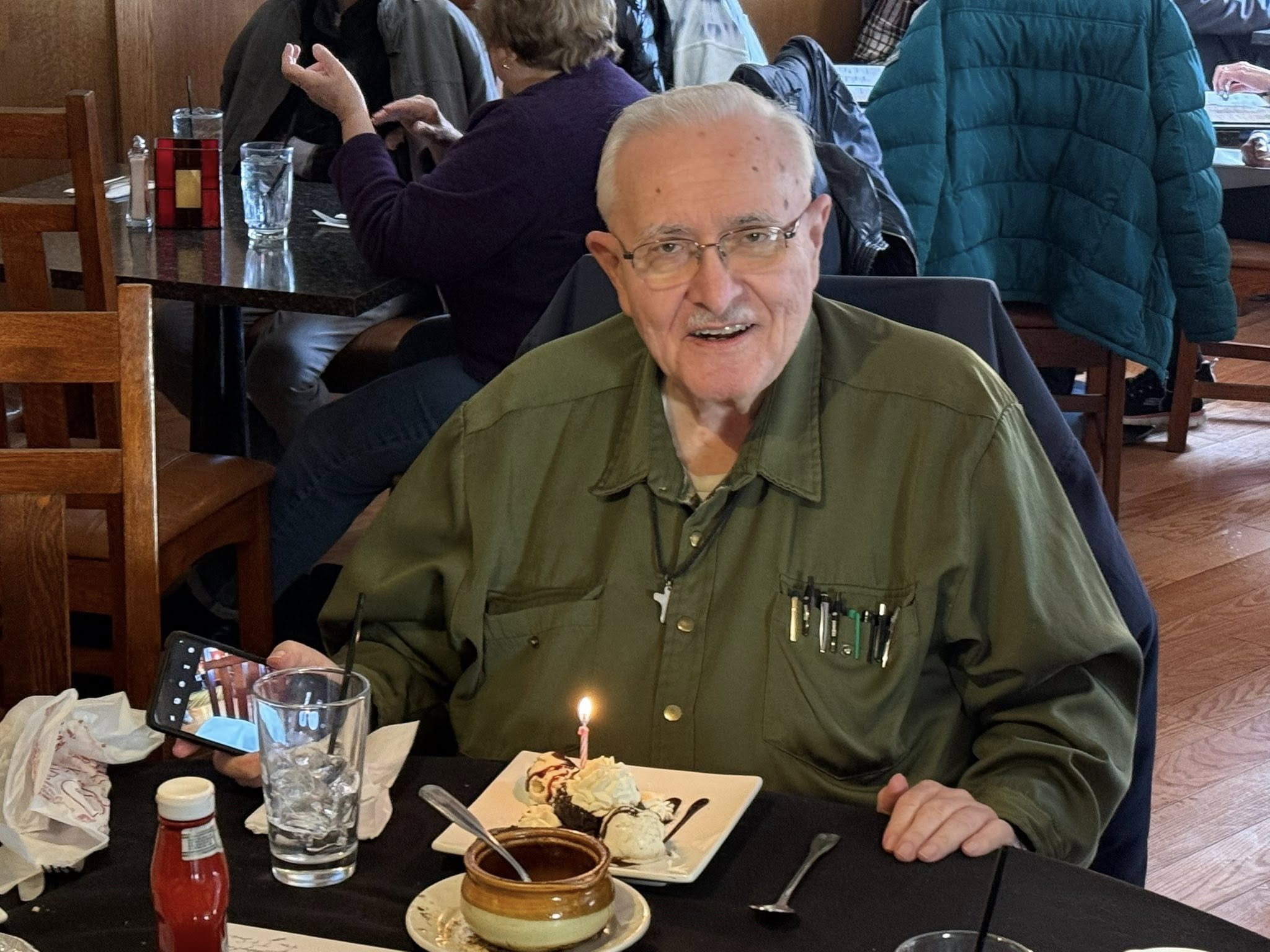 An elderly man with glasses and a mustache, wearing a green shirt with pens in the pocket, sitting at a restaurant table with a birthday dessert with a lit candle, holding a smartphone in his left hand, and smiling at the camera.