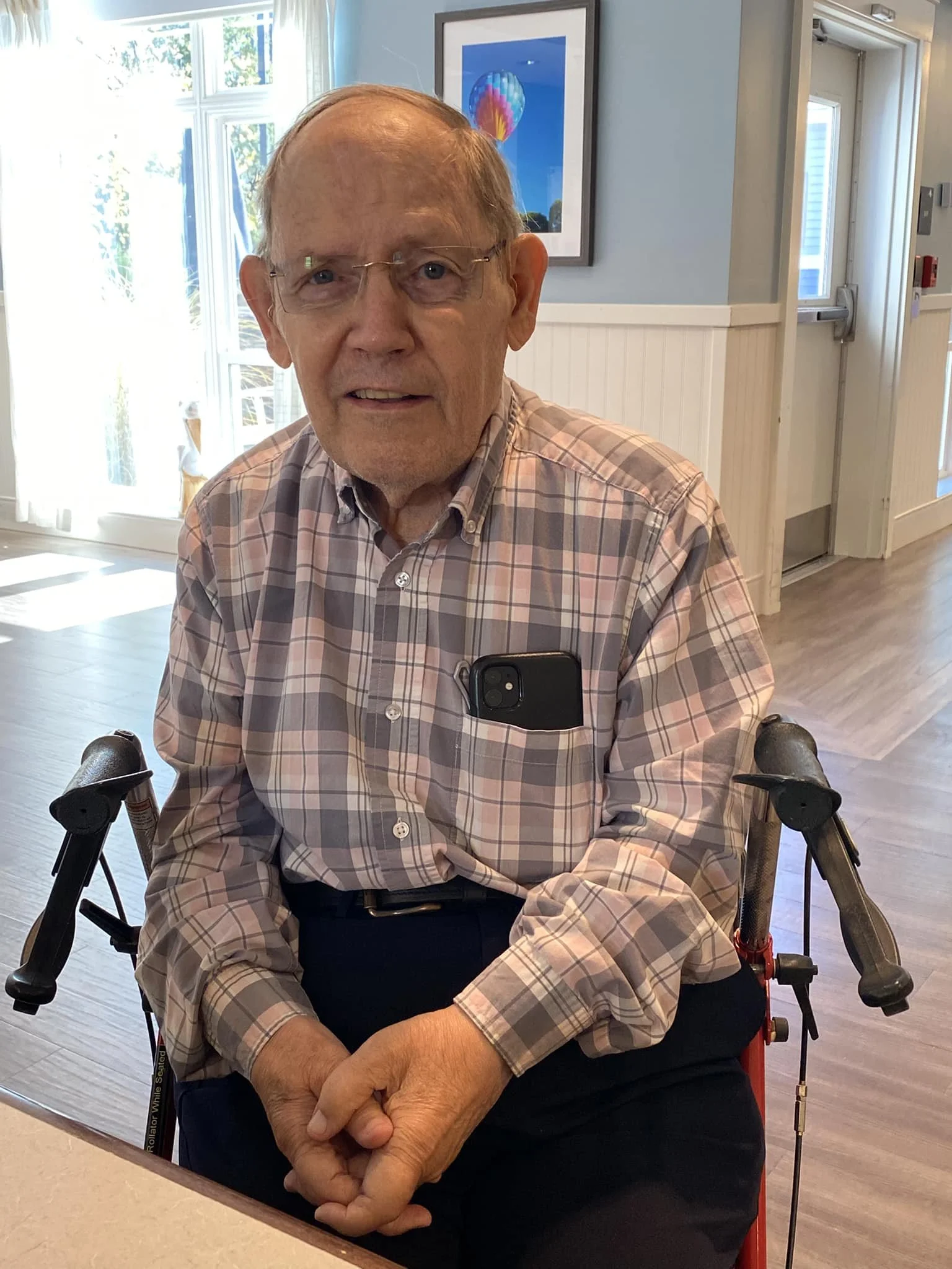 An elderly man sitting in a wheelchair, wearing glasses and a plaid shirt, indoors with a framed artwork and windows in the background.