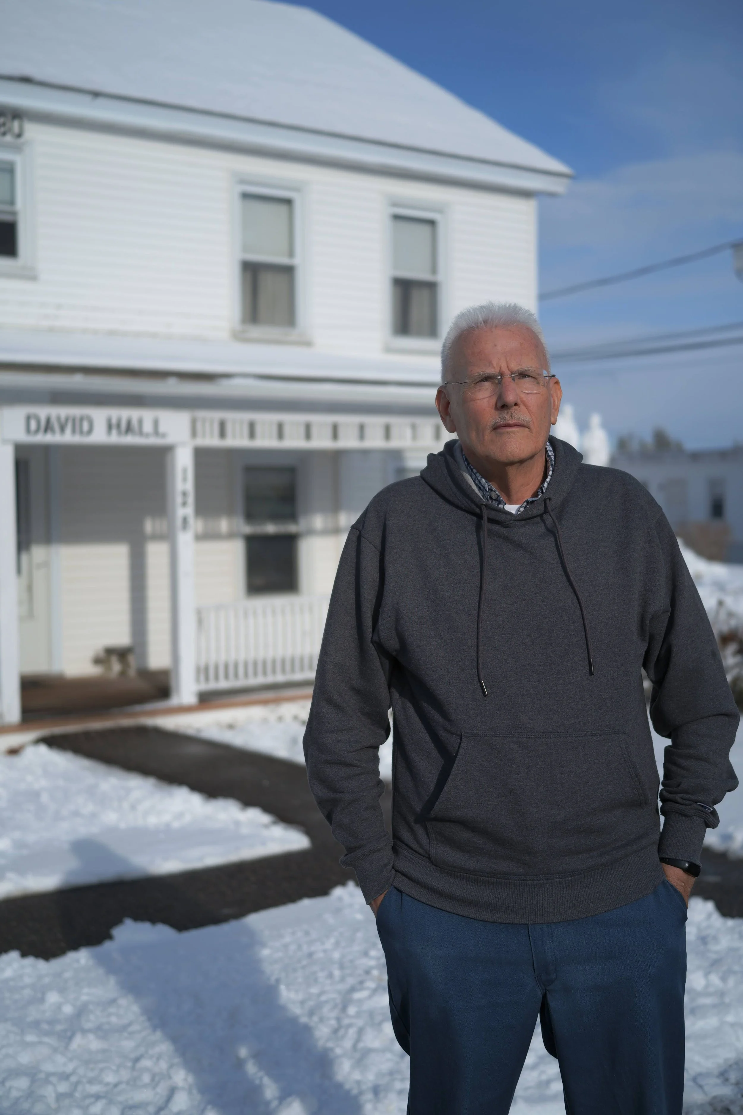 An elderly man with white hair and glasses stands outside on a snowy sidewalk in front of a white house, wearing a dark hoodie and blue pants.