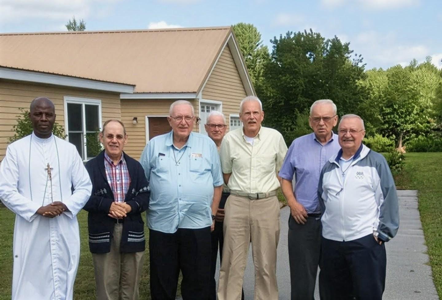 Seven men standing outside in front of a house with a beige exterior and green trees, some wearing glasses and casual or religious attire.