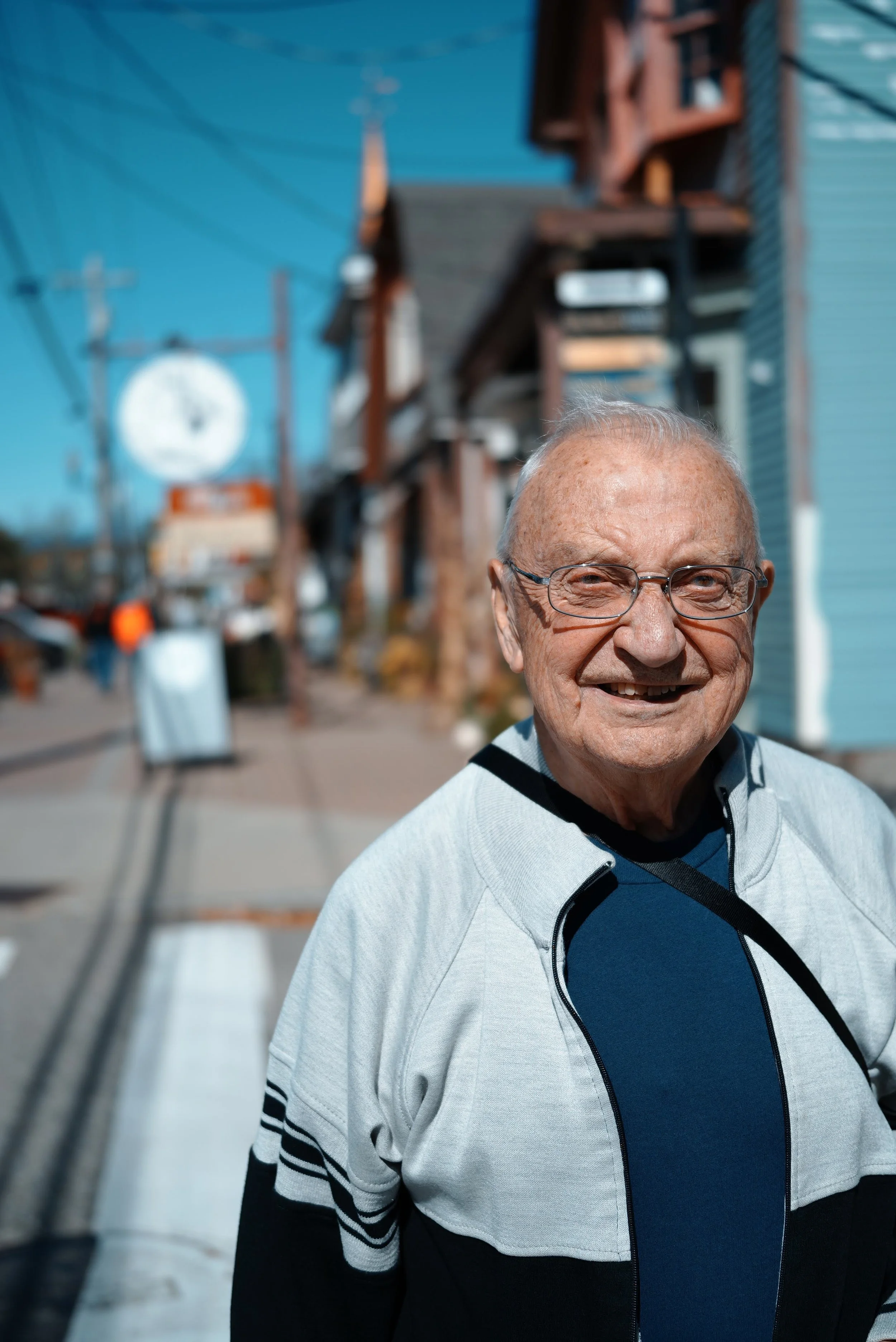 An elderly man wearing glasses and a light-colored zip-up jacket is smiling at the camera on a city street with bright blue buildings in the background.