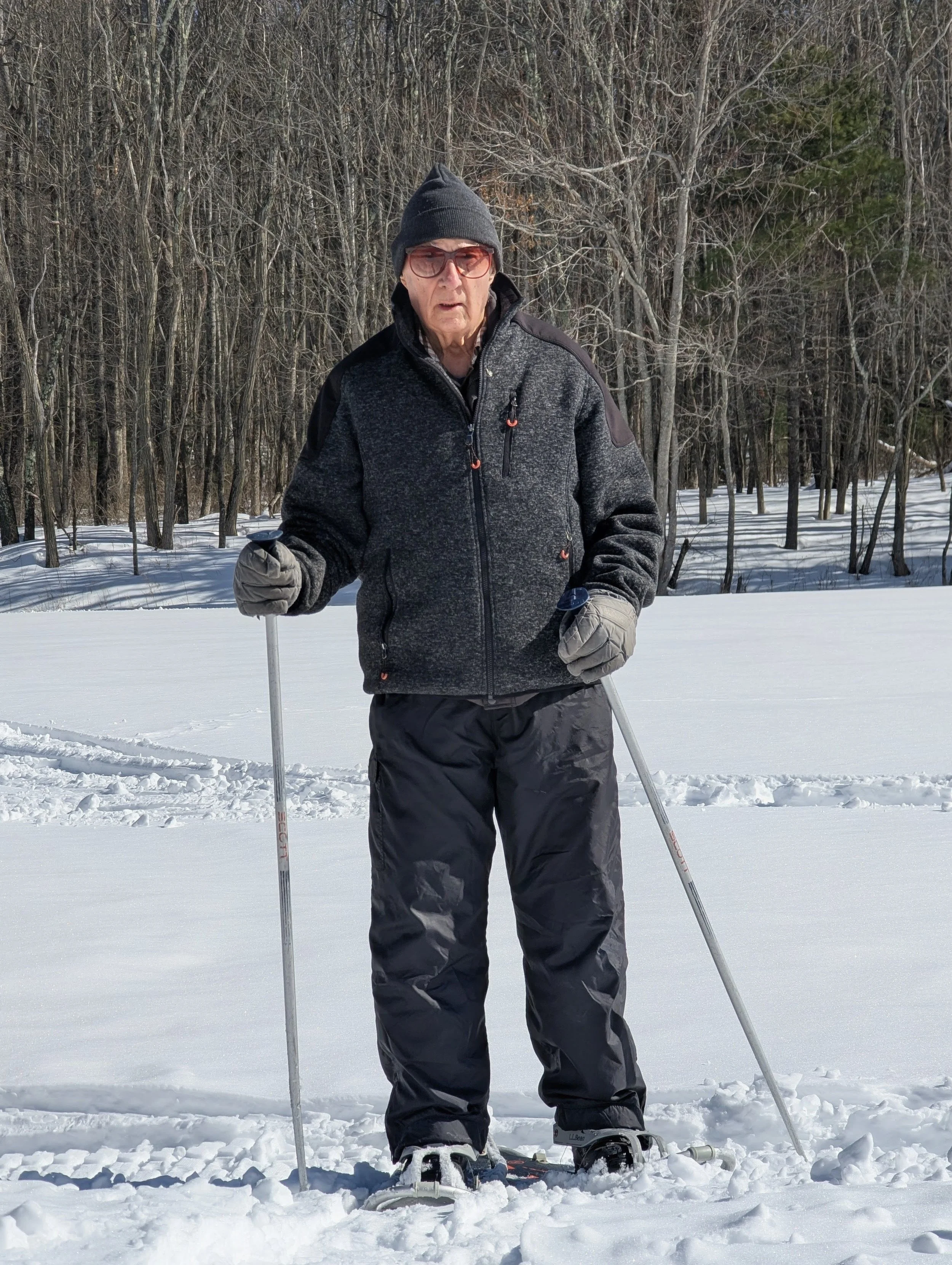 An elderly person wearing winter gear, including a dark beanie, glasses, gloves, a black jacket, and black pants, standing on snow with ski poles and skis, in a snowy, wooded area.