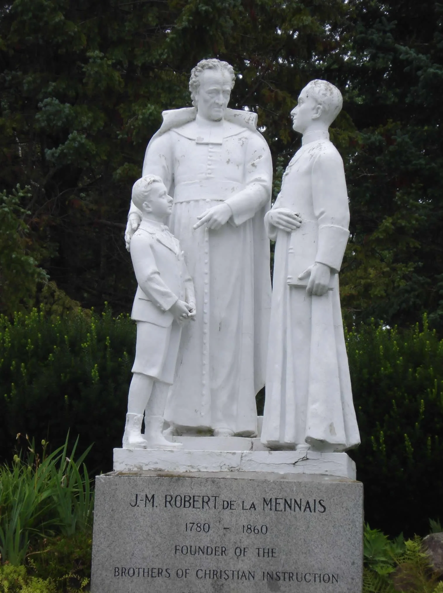 White statue of a man, woman, and child standing outdoors, with trees in the background. The man appears to be explaining something to the woman and child. The statue's base has an inscription that reads "J-M ROBERT de LA MENNAIS 1780-1860 FOUNDER OF THE BROTHERS OF CHRISTIAN INSTRUCTION".