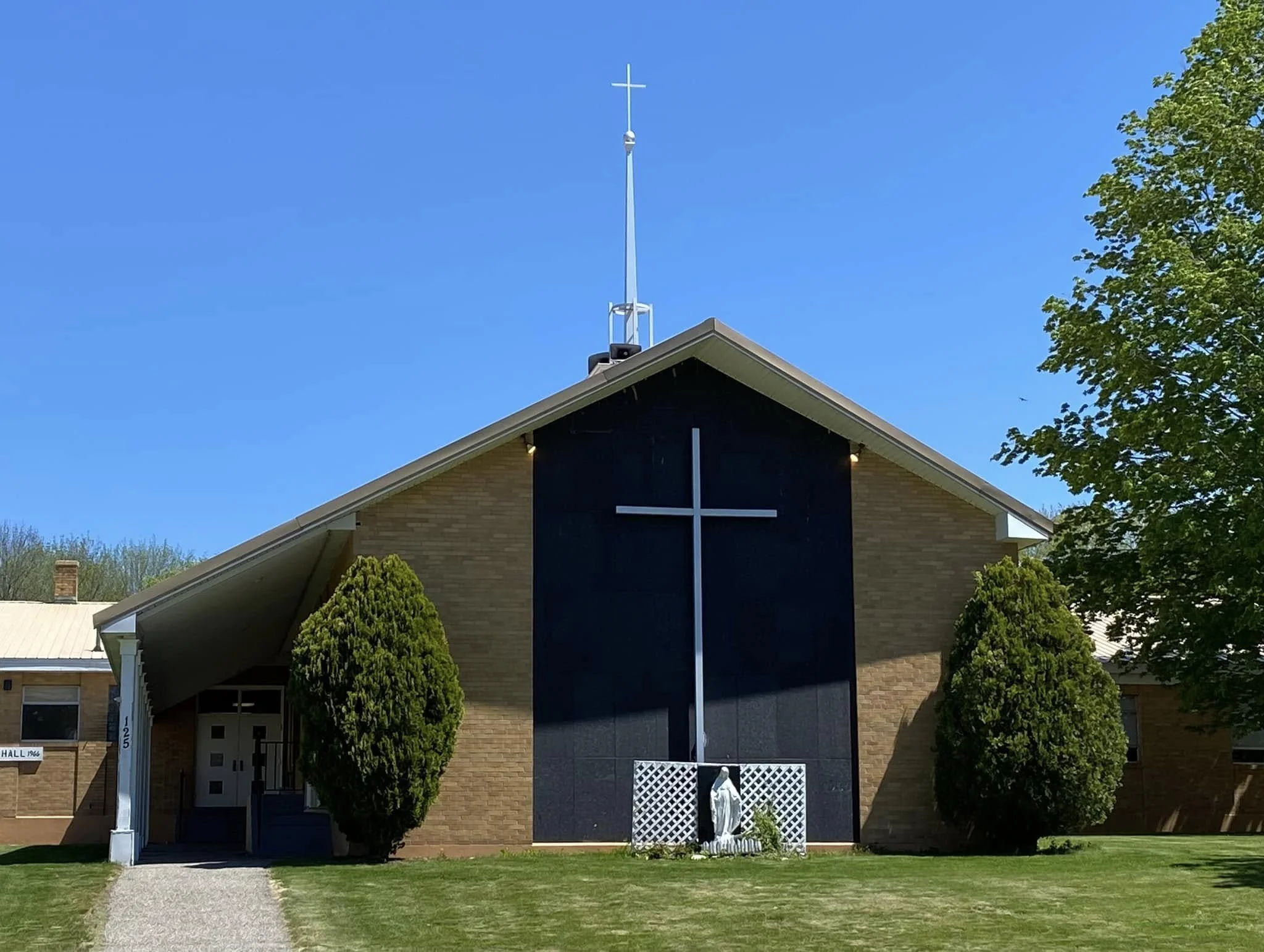 Front view of a church building with a large cross on the front. The church has a sloped roof, brick walls, and two symmetrical bushes on either side of a religious statue in front. Blue sky and part of a tree are visible.