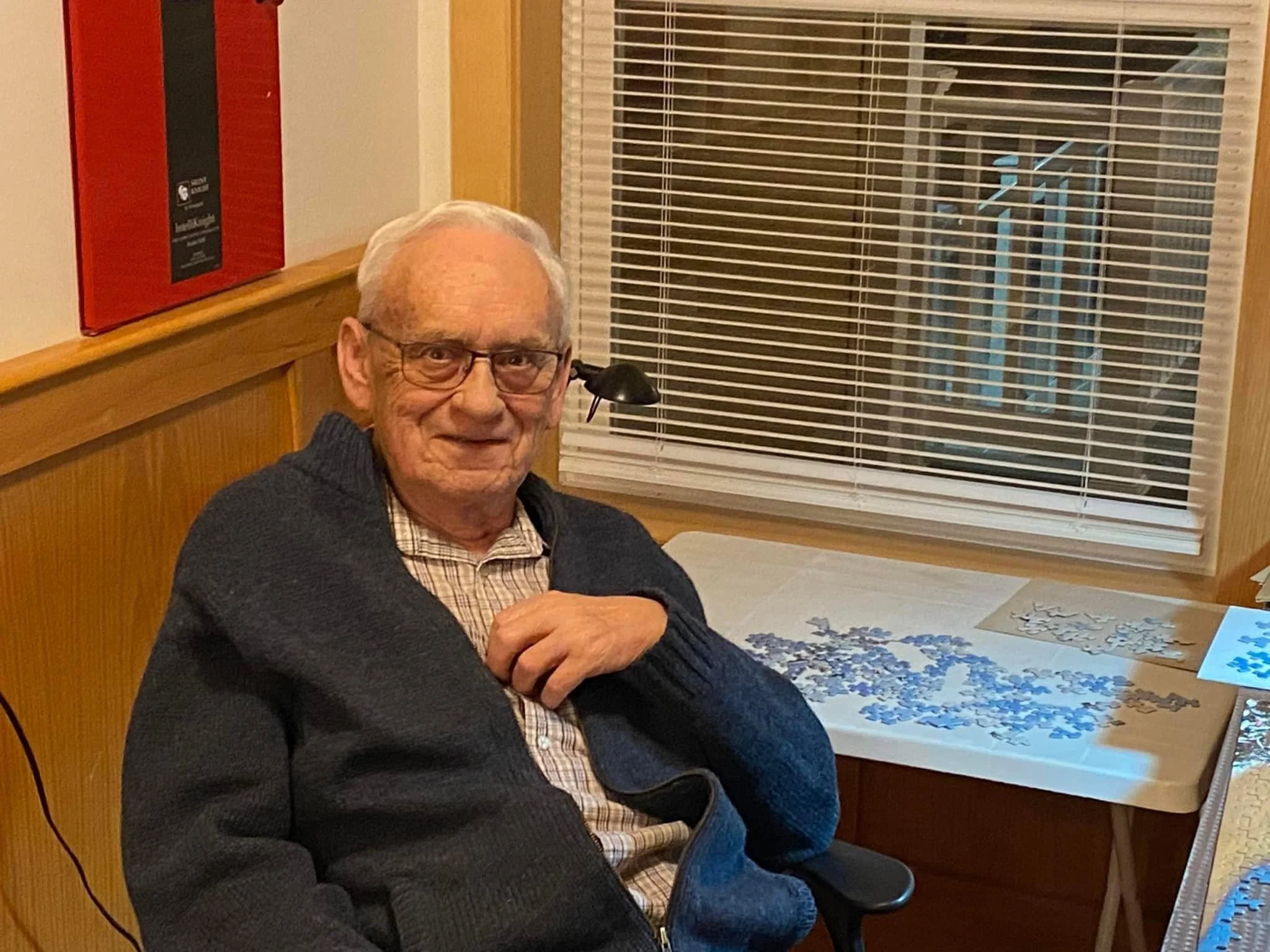 An elderly man with glasses, white hair, and a checkered shirt sitting at a table with scattered puzzles, in a room with wood-paneled walls and a window with closed blinds.