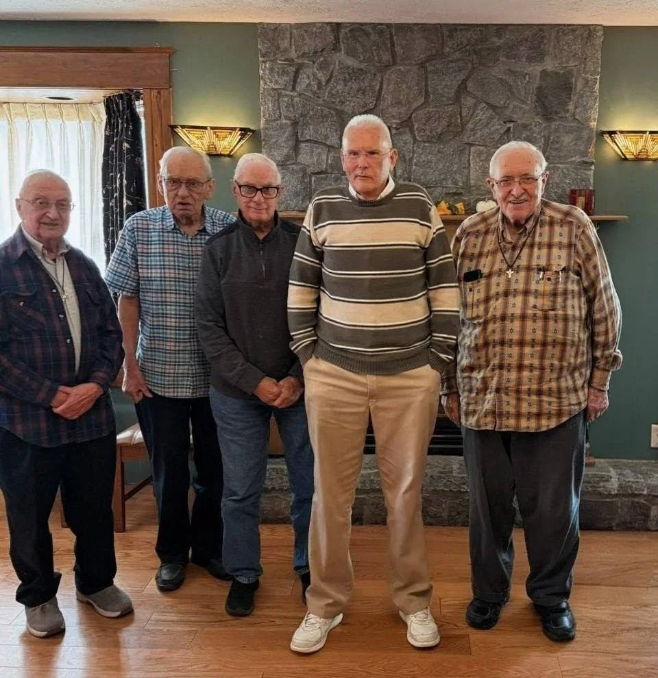 Five elderly men standing in a room with a stone fireplace, wooden flooring, and window with curtains, posing for a photo.