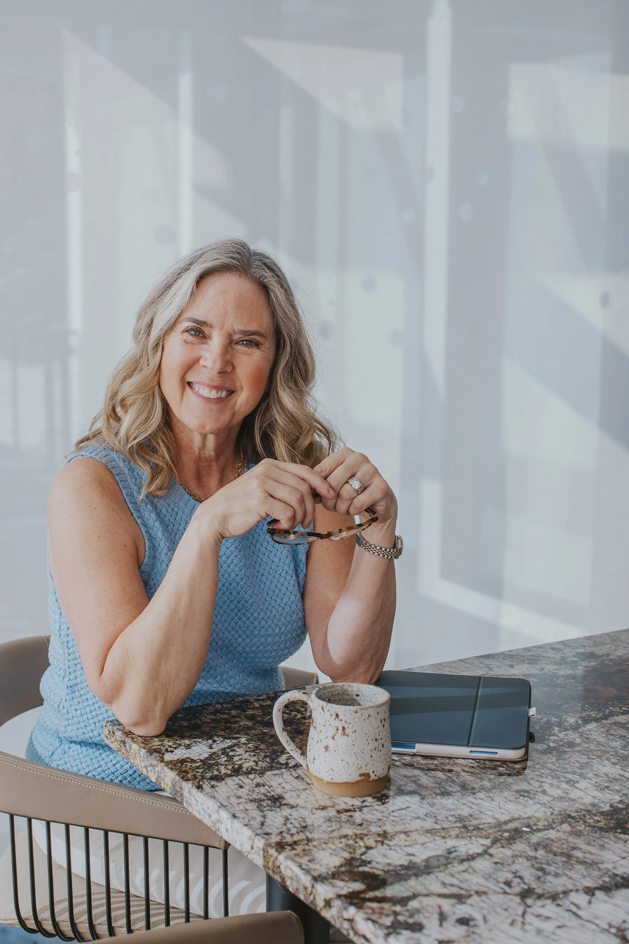 A smiling woman with blonde-gray hair in a blue sleeveless top sits at a marble table with a coffee mug, holding glasses
