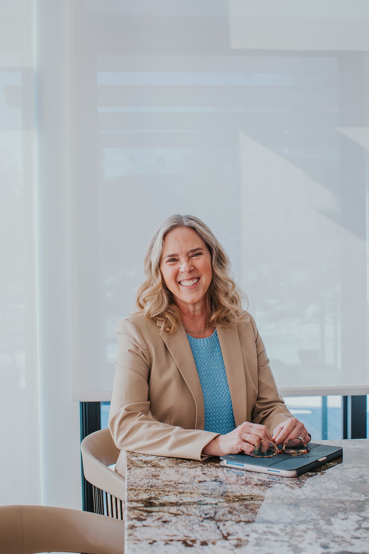 A smiling woman with blonde-gray hair in a tan blazer and blue top sits at a marble table, holding glasses