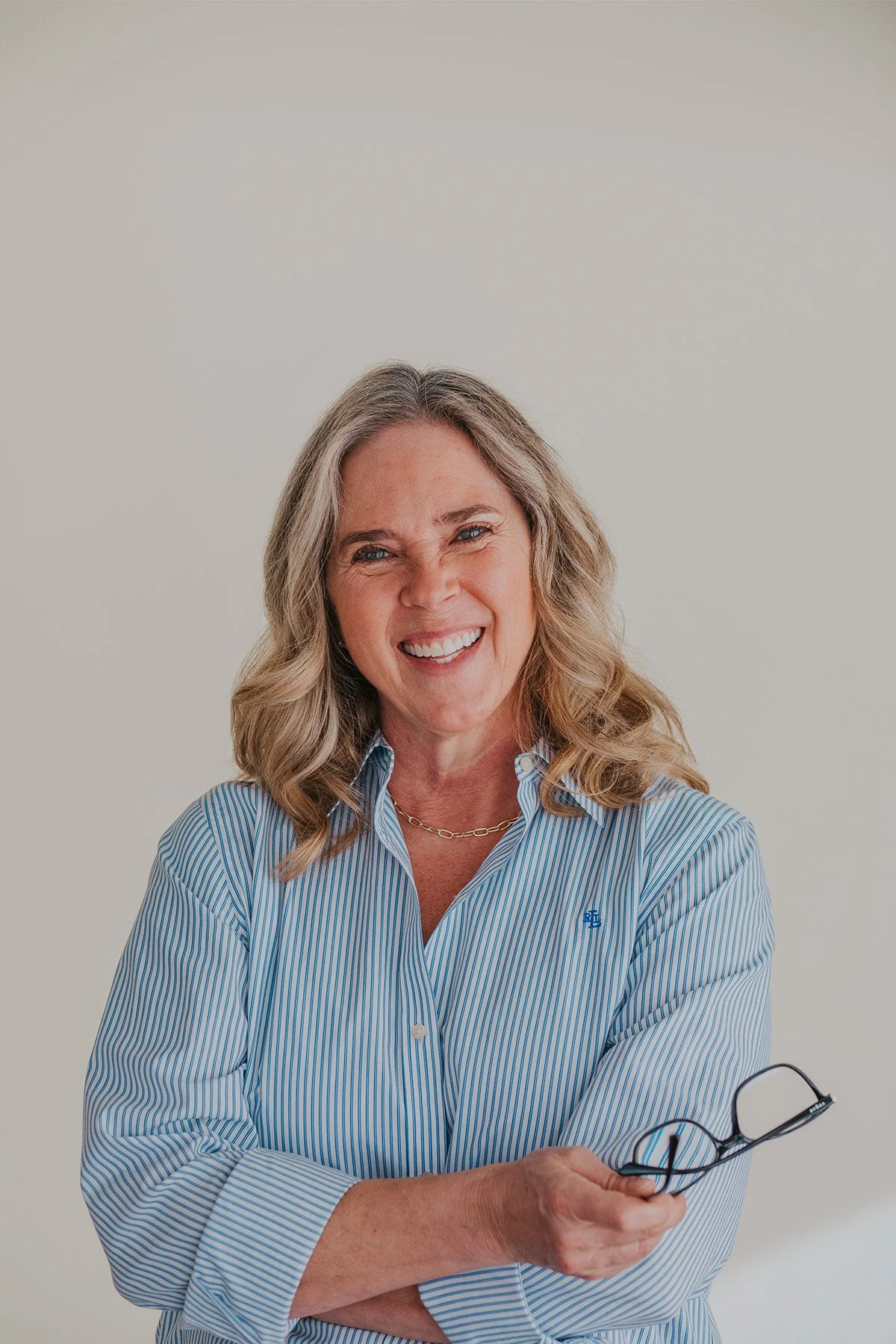 A smiling woman with blonde-gray hair in a blue striped shirt stands with arms crossed, holding glasses