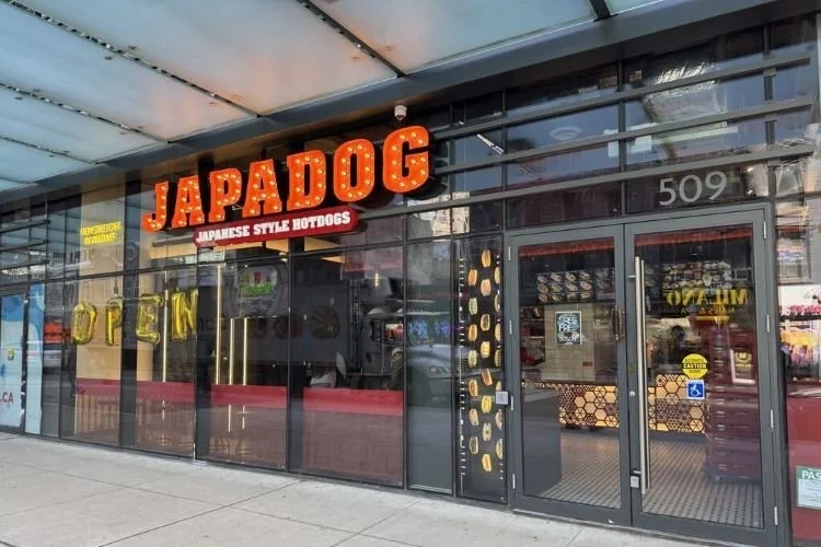The storefront of Japanese style hotdog restaurant named JAPADOG with large glass windows, a red sign with white and red letters, and an open sign in the window.