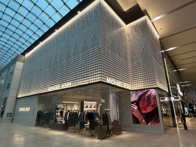 Exterior of a Michael Kors store inside a modern shopping mall with glass ceiling and large digital advertisement featuring a woman.