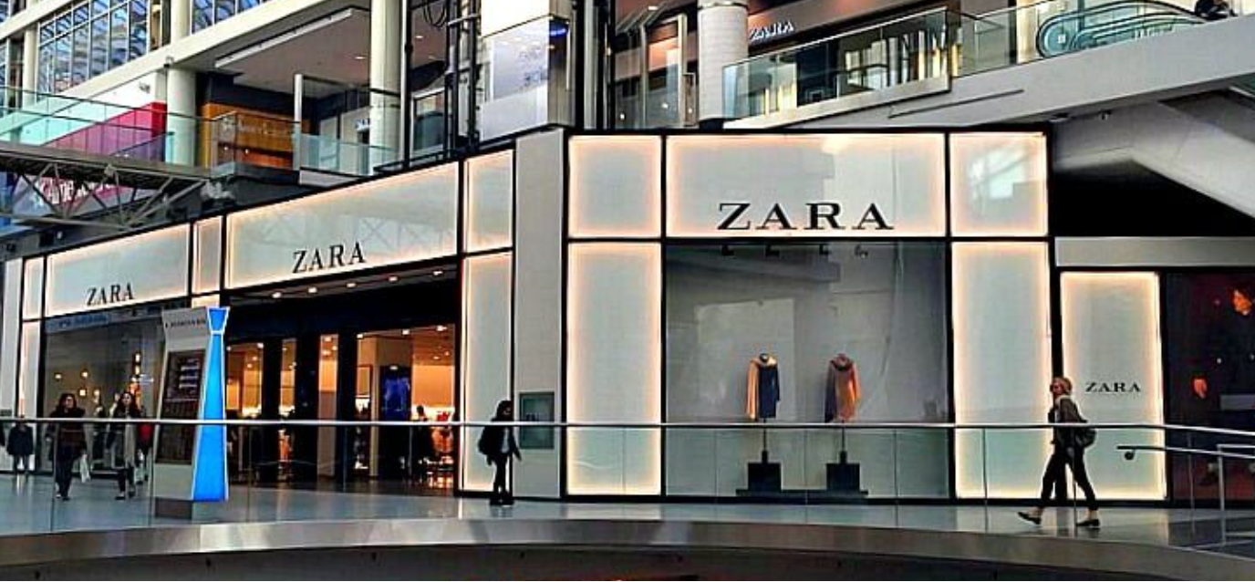 Exterior view of a Zara clothing store inside a shopping mall, with illuminated signage and window displays of mannequins dressed in clothing.
