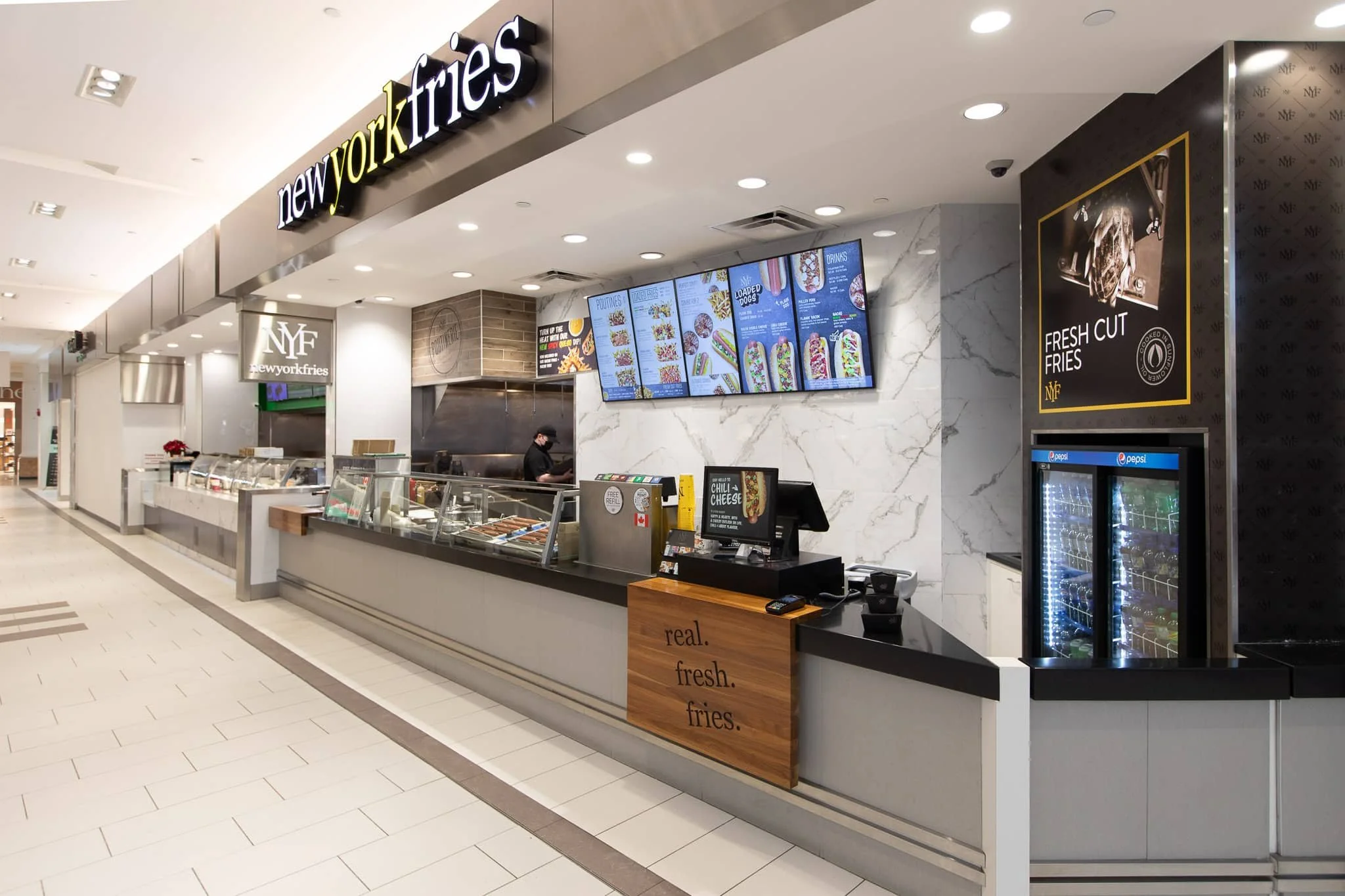A food kiosk named "new york fries" inside a shopping mall. The kiosk offers fries, hot dogs, poutine, and other snacks, with digital menu screens displaying the options. There's a small refrigerator with beverages beside the counter, and a worker preparing orders behind the counter.