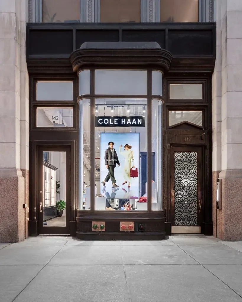 Storefront with large glass display window showcasing mannequins and a digital advertisement, branded 'COLE HAAN', black trim, and a decorative black door to the right, located at 145 on the street.