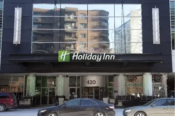The exterior of a Holiday Inn hotel with large glass windows, the Holiday Inn logo, and the street number 420, with parked cars in front.