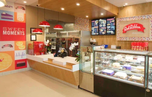 Inside a Red Ribbon bakery shop with display case of cakes and desserts, decorated walls, and menu boards behind the counter.