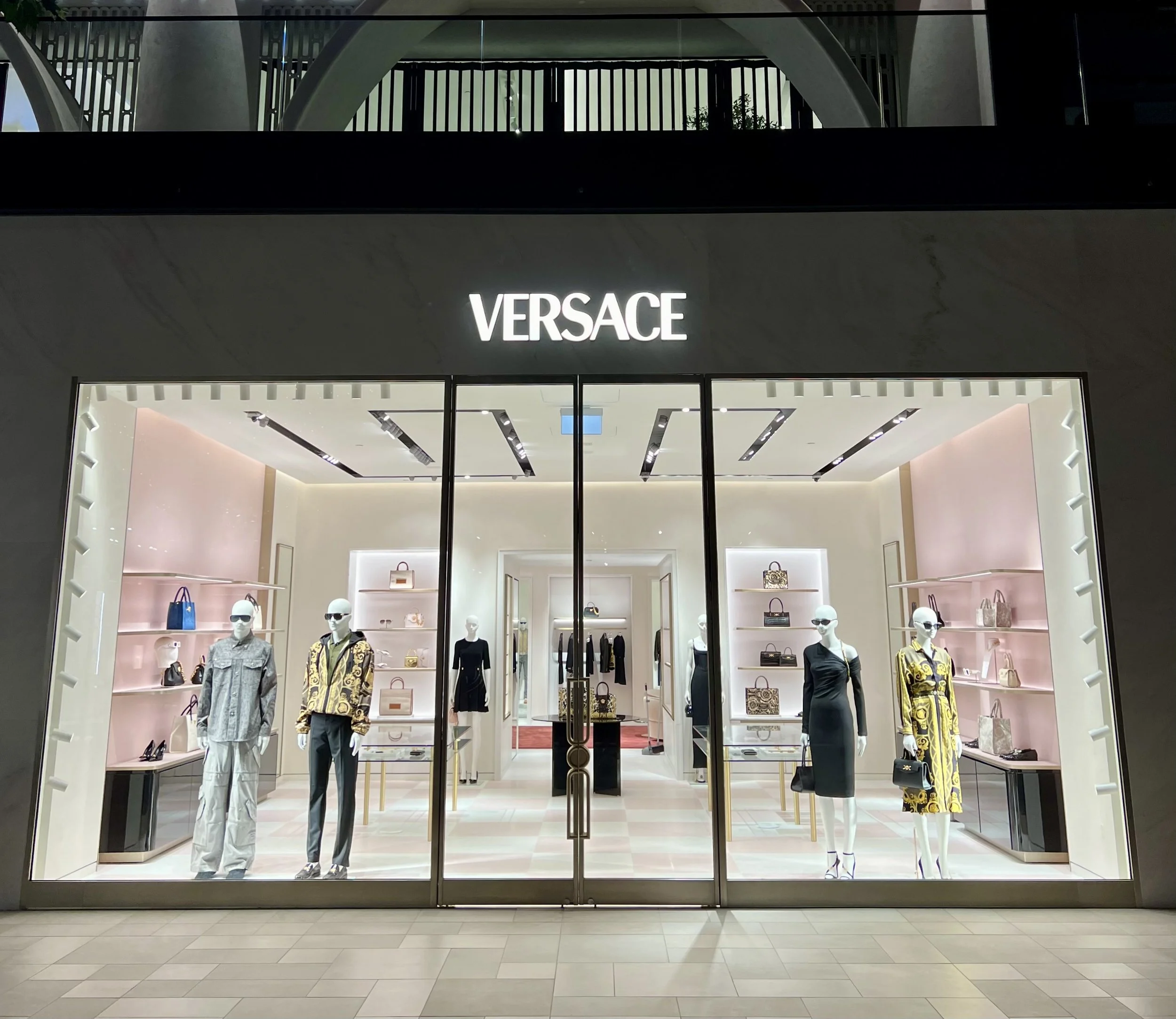 The exterior of a Versace store at night with mannequins in the window displaying fashionable clothing and accessories, illuminated signage, and a modern storefront design.