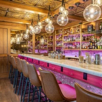 A stylish bar with a marble countertop, brown bar stools, and shelves stocked with various bottles, illuminated by hanging globe lights and purple accent lighting.