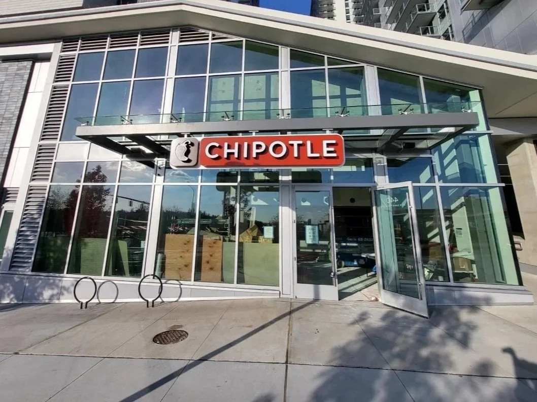 Exterior view of a modern Chipotle restaurant with large glass windows, a metal awning, and a prominent red and white Chipotle sign above the entrance.