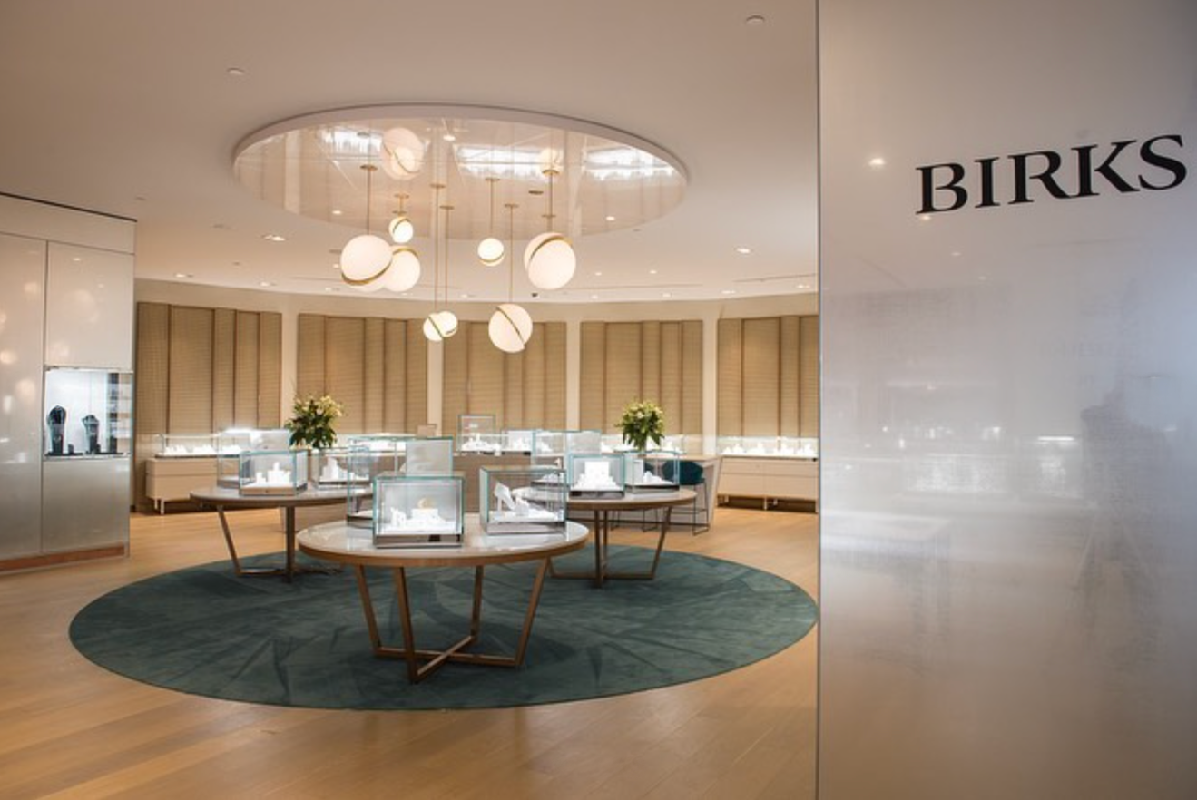 Inside Birks jewelry store with display cases and modern hanging lights, featuring a circular green rug on wooden flooring.