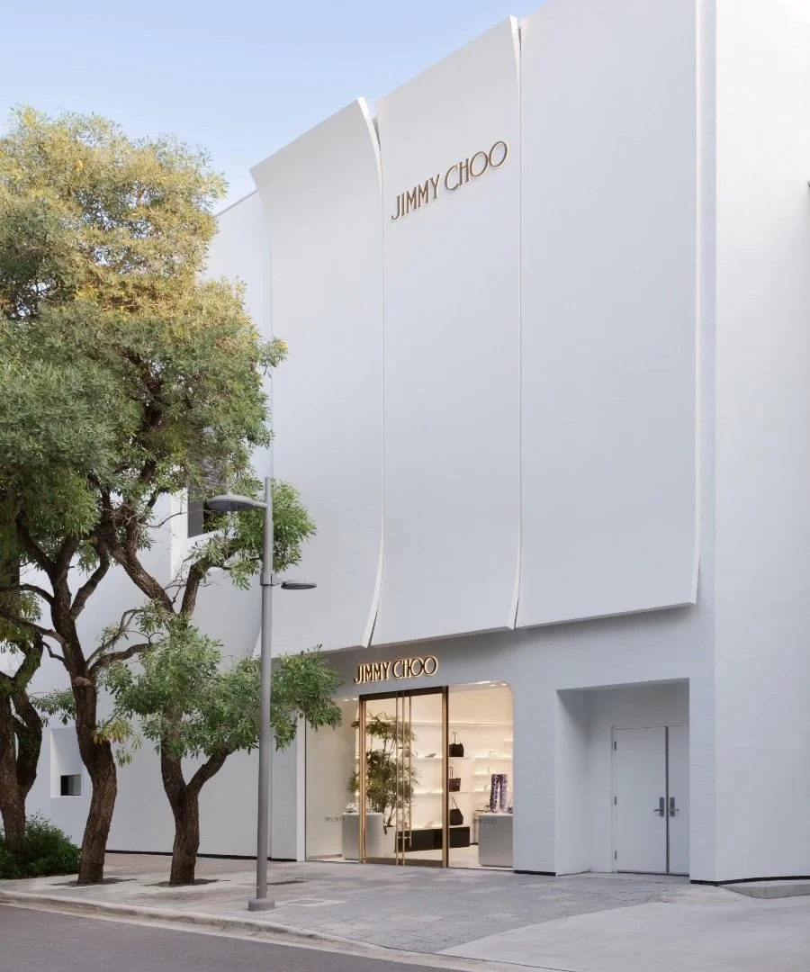 Exterior view of a Jimmy Choo store with a white modern facade, large signage, and glass entrance doors. There are trees in front of the store and street lamps.