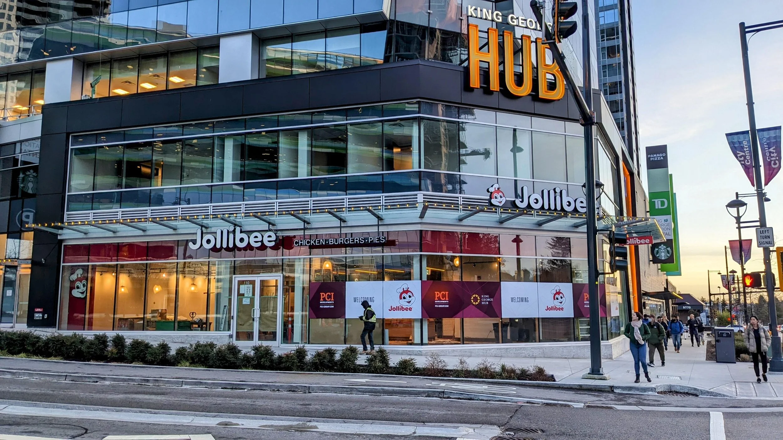 A corner storefront of a modern glass building with signs for Jollibee and King George Hub, with people walking along the sidewalk and a person in safety gear near the entrance.