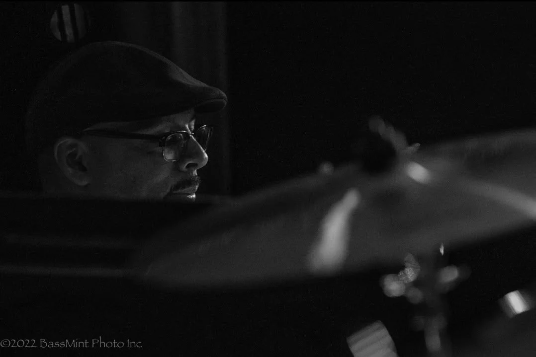 A man wearing glasses and a flat cap, seen in profile, appears to be playing the drums in a dark room.