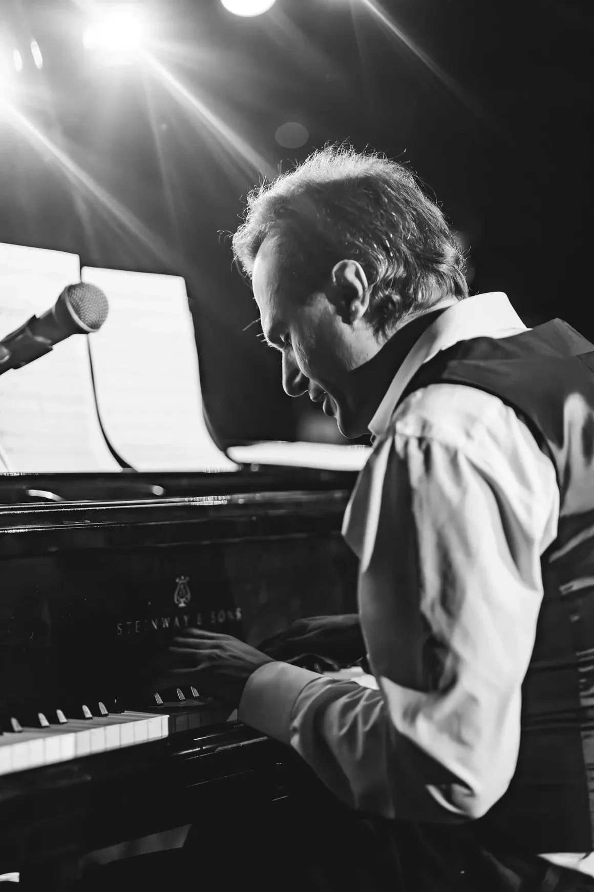 Black and white photo of a man playing a grand piano with a microphone nearby, illuminated by stage lights.
