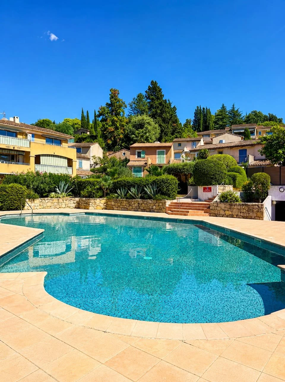Une piscine extérieure claire avec une zone de repos en pierre, entourée de végétation luxuriante et de maisons colorées sur une colline, sous un ciel bleu sans nuages.