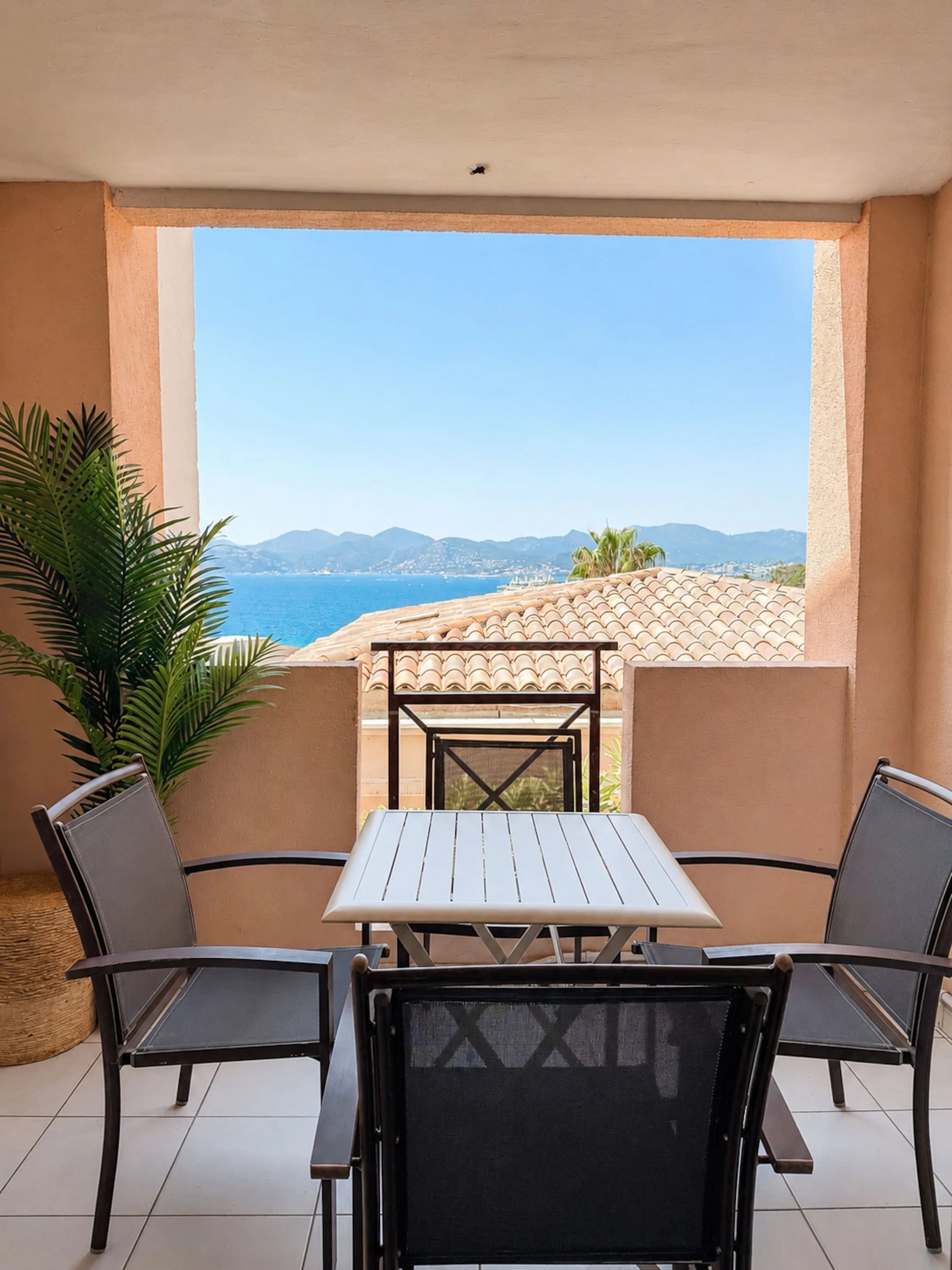 Balcon avec table et chaises en vue vers la mer, montagnes en arrière-plan, ciel clair, végétation luxuriante