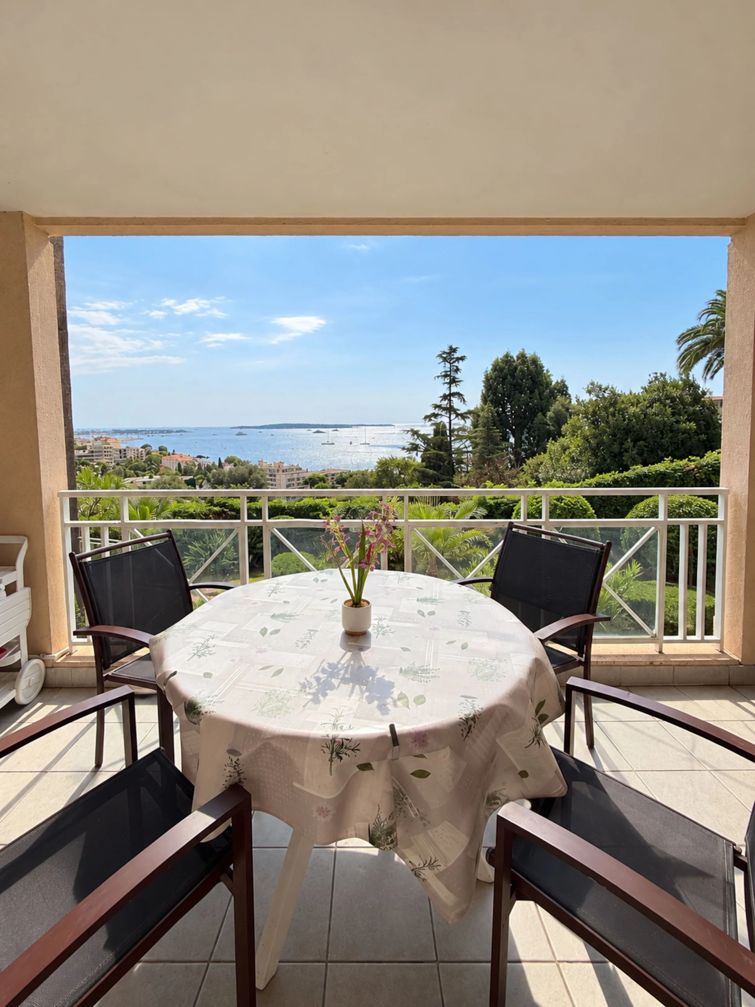 Vue depuis une terrasse avec une table ronde avec une nappe à motifs floraux, cinq chaises en bois avec assises en tissu noir, un petit vase avec des fleurs au centre, et un paysage avec la mer, des arbres, un ciel bleu et quelques nuages en arrière-plan.