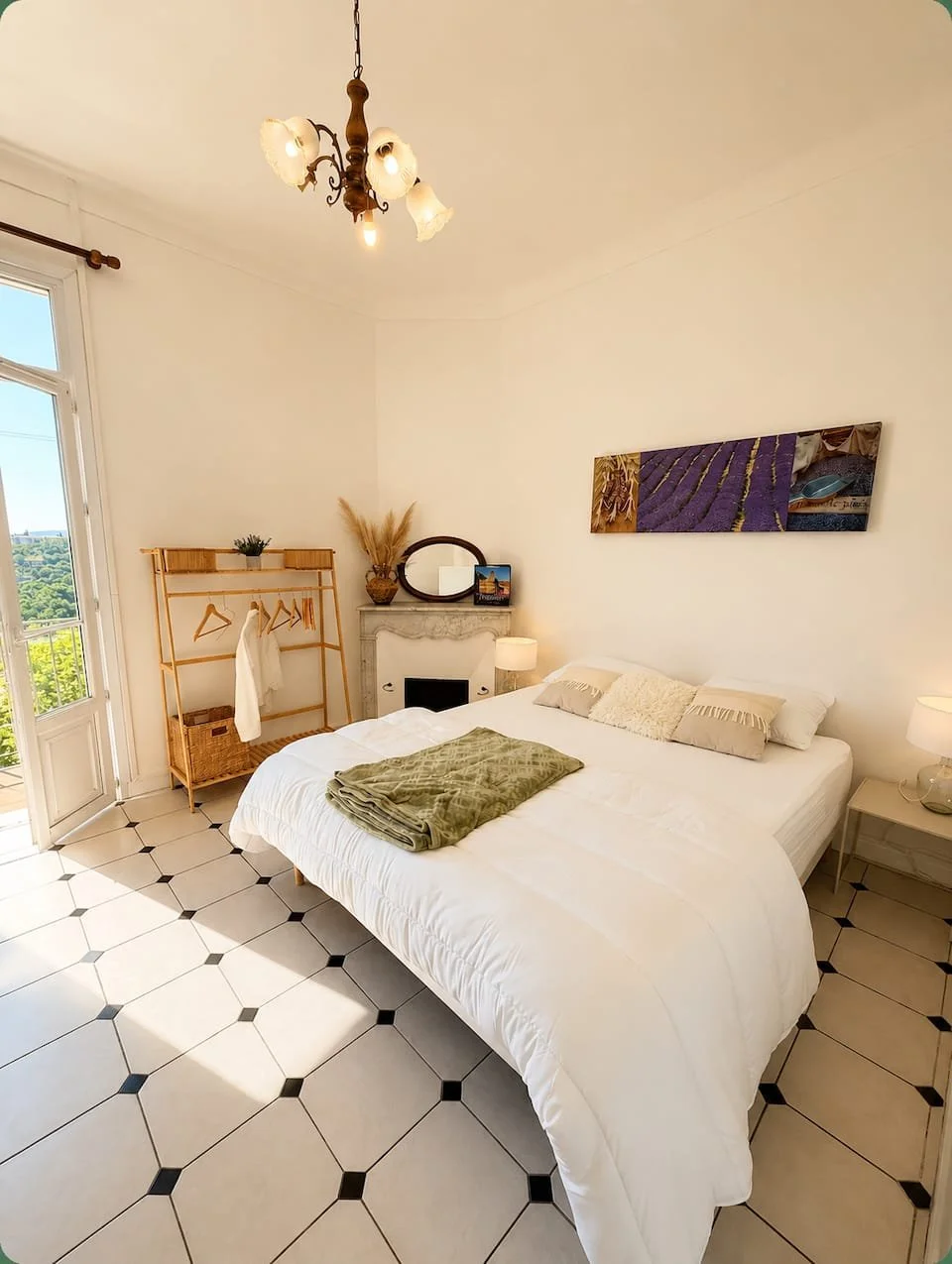 Chambre lumineuse avec un grand lit blanc, une cheminée en marbre, deux tables de nuit avec lampes, un tapis vert, une penderie en bois, une porte en verre ouverte sur un balcon avec vue sur la nature, un tableau de lavande, un chandelier suspendu au plafond.