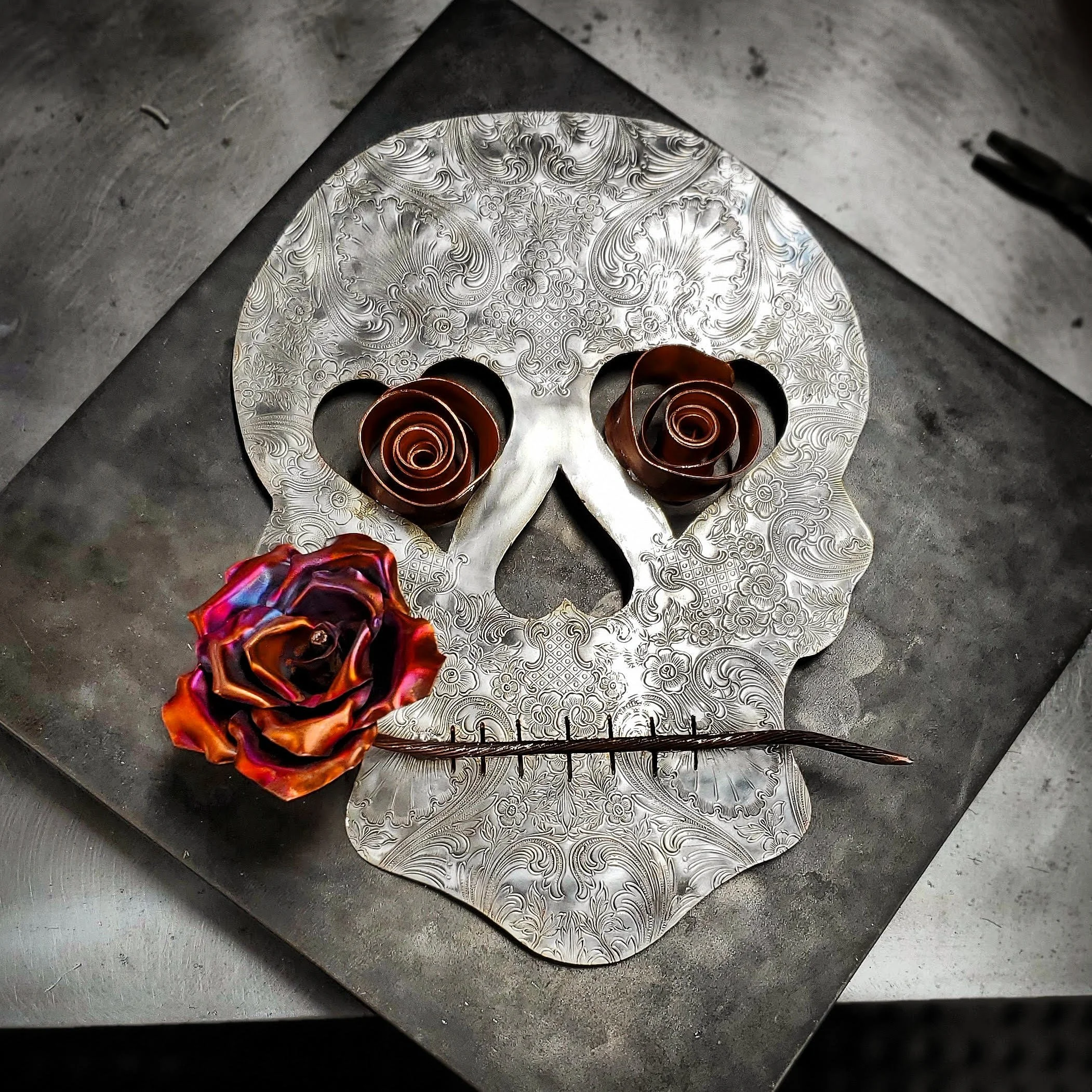Decorative metal skull with intricate engraved patterns, adorned with three spiral metal roses and a metallic rose in the lower left, on a black surface.
