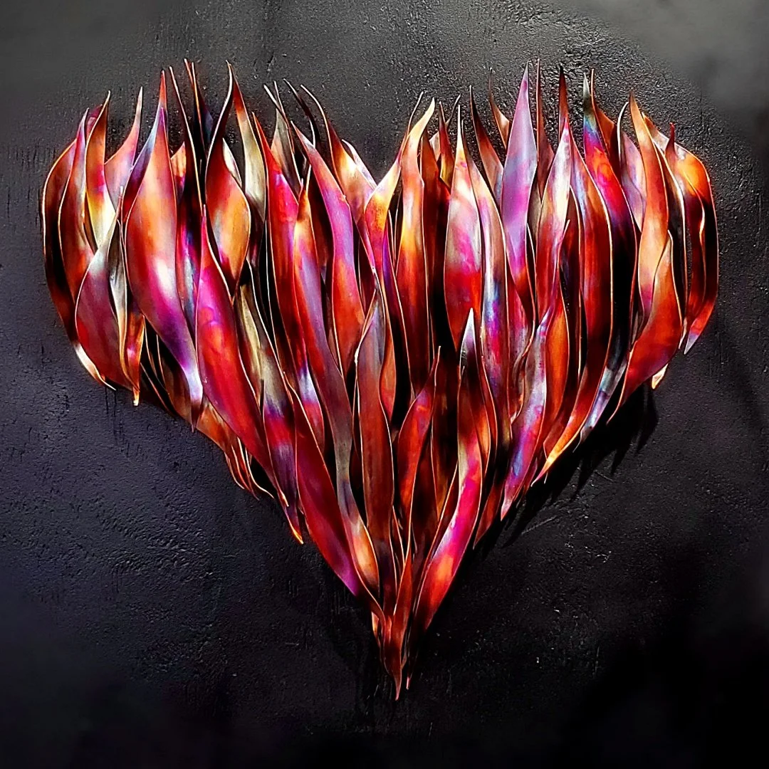 Flame-painted copper heart wall art sculpture with uneven flame shaped edges. It has a red finish with the shifting colors of fire and a shiny surface