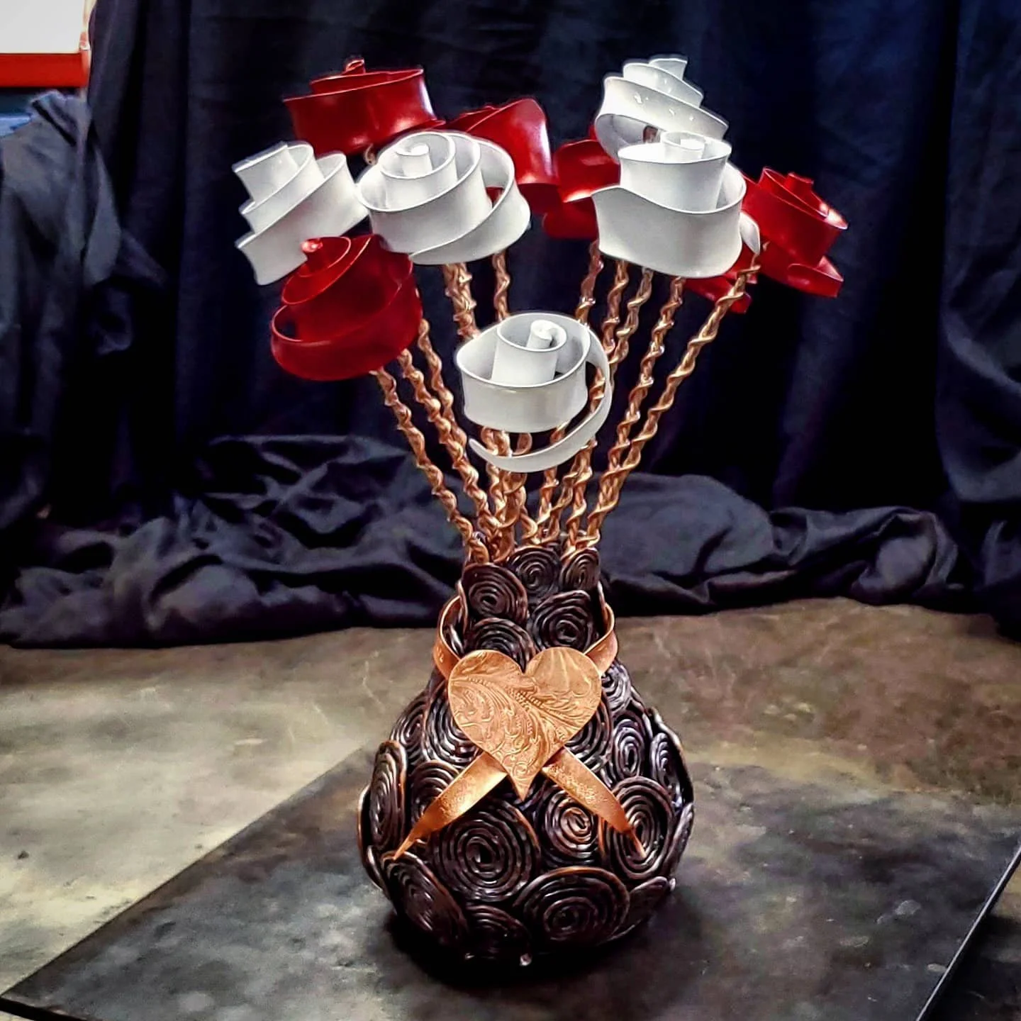 Decorative vase made of swirling black and brown metal with a copper heart and arrow ornament wrapped around it. The vase has curly metal rods extending upward, topped with red and white metal roses. The arrangement is set on a dark surface with a black backdrop.