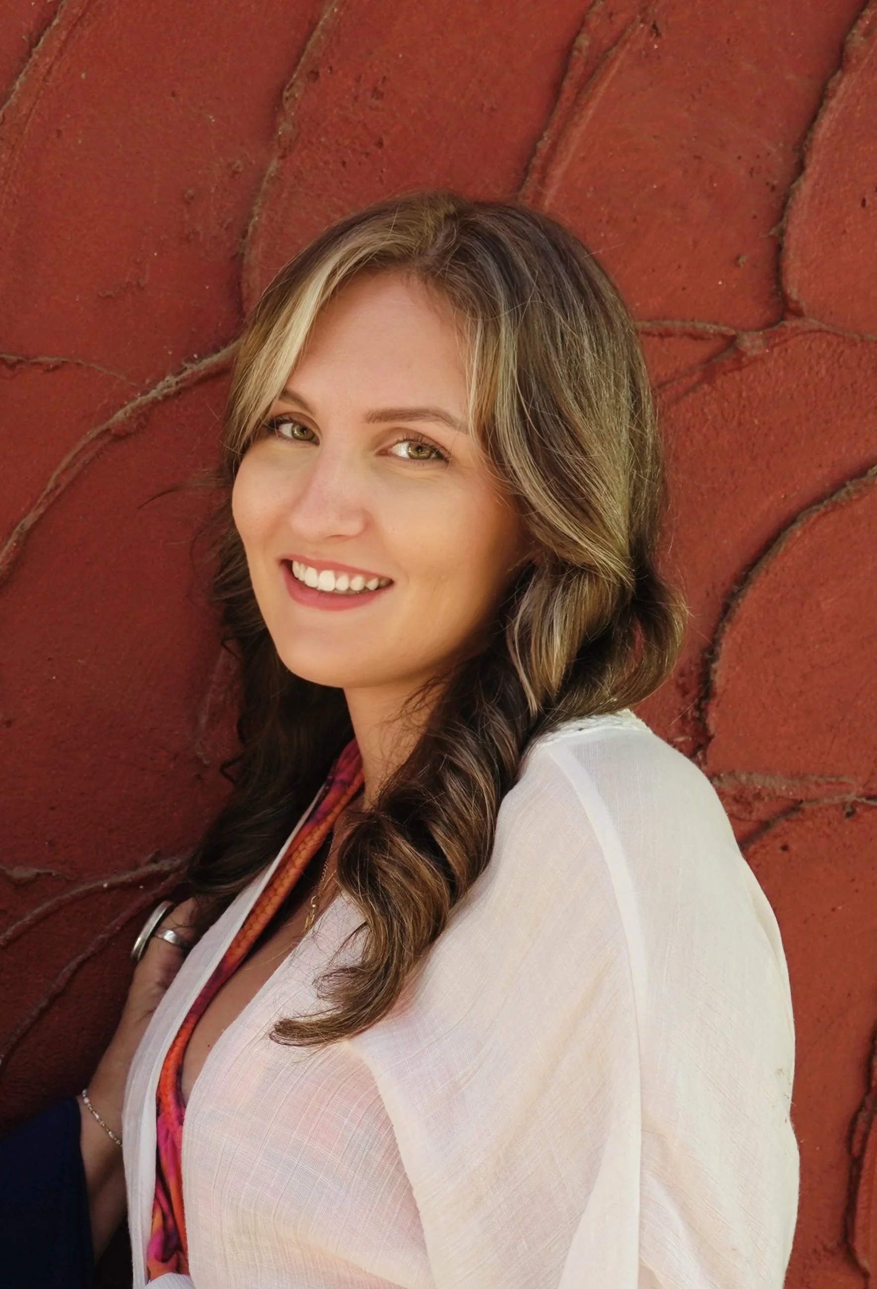 A woman with long wavy brown hair and light green eyes smiling and looking to the side, standing against a red brick wall.