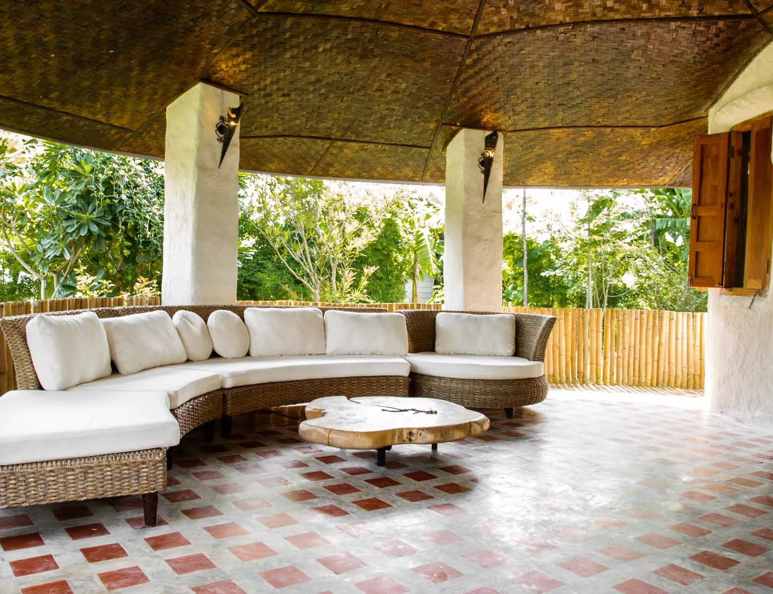 Covered outdoor patio area with a curved white cushioned wicker sofa, a wooden coffee table, potted plants, and open shutters, surrounded by greenery.