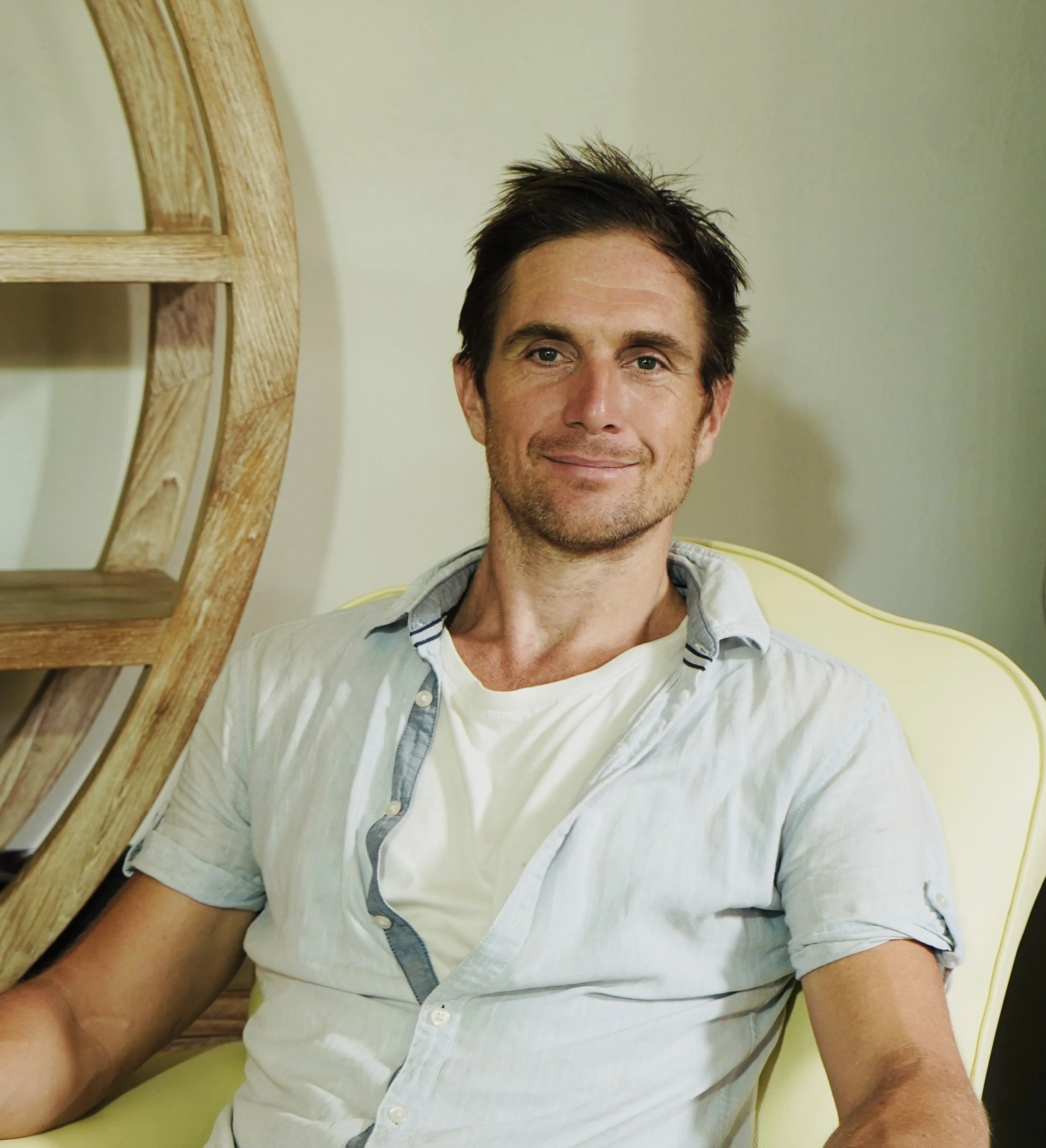 A man with short dark hair and light stubble smiling, sitting on a yellow chair next to a wooden ladder, wearing a light-colored, button-up shirt with rolled sleeves, against a plain beige wall.