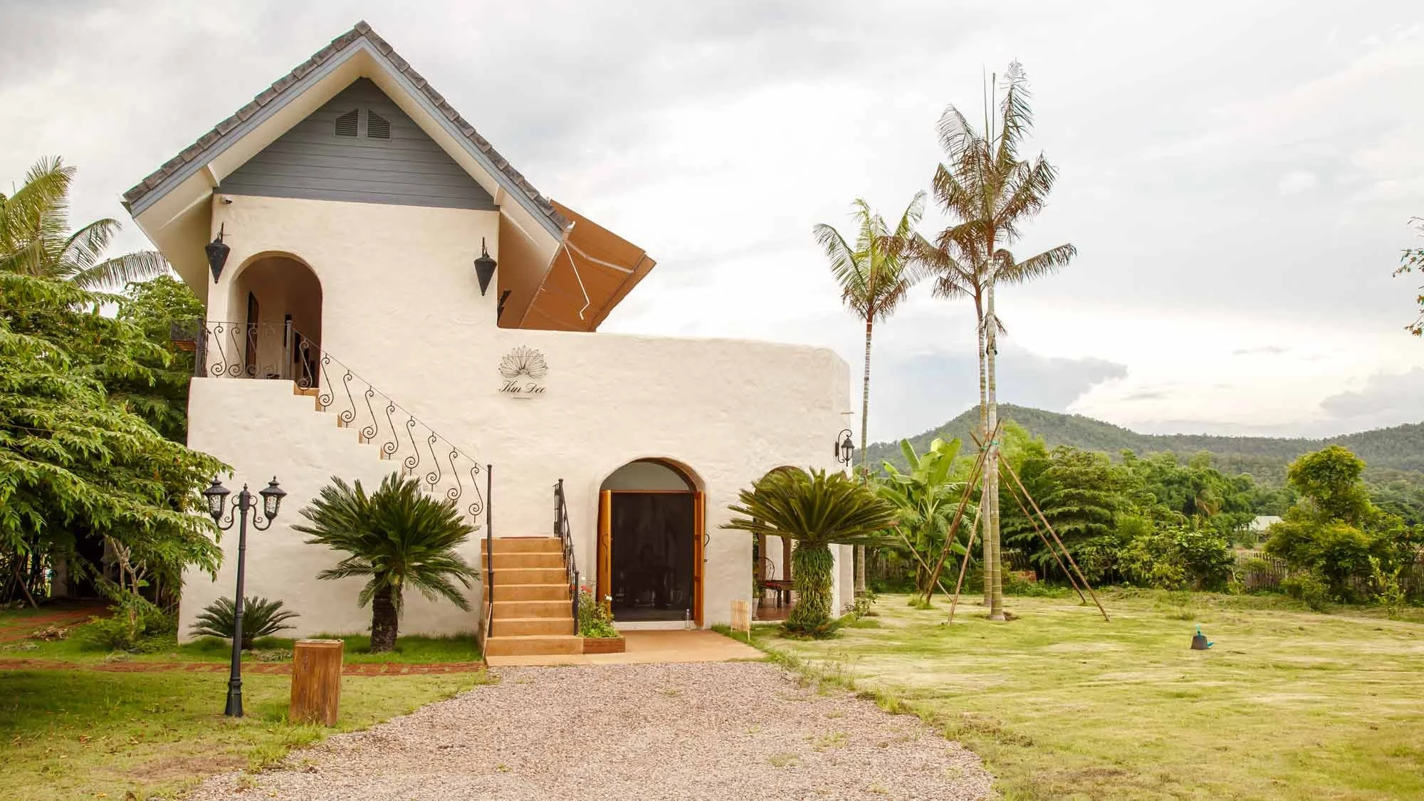 A white two-story house with a gray roof and a small front porch, surrounded by lush greenery, trees, and a mountain in the background.